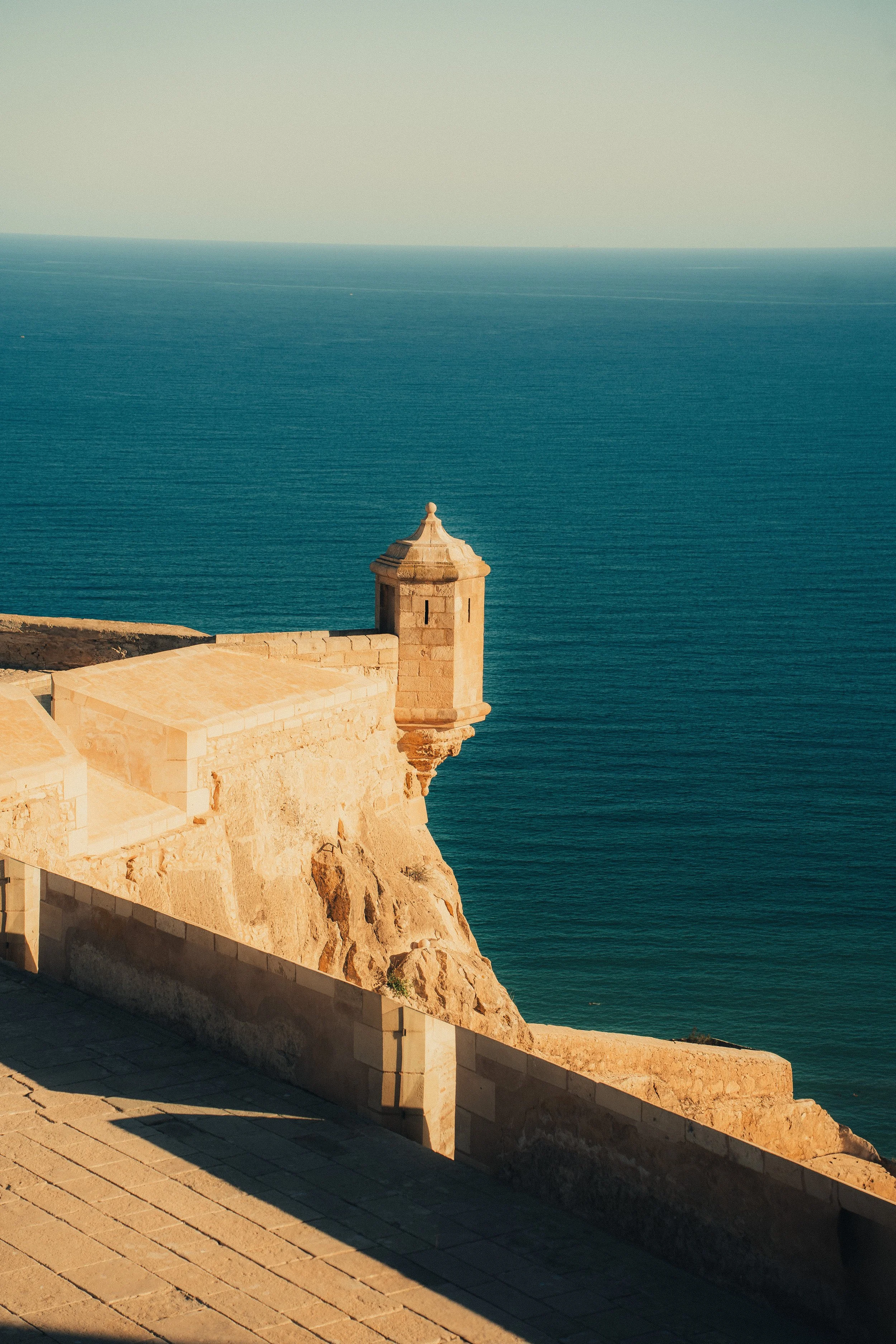 A stone fortress overlooking the ocean, featuring a small turret on a cliffside at sunset.