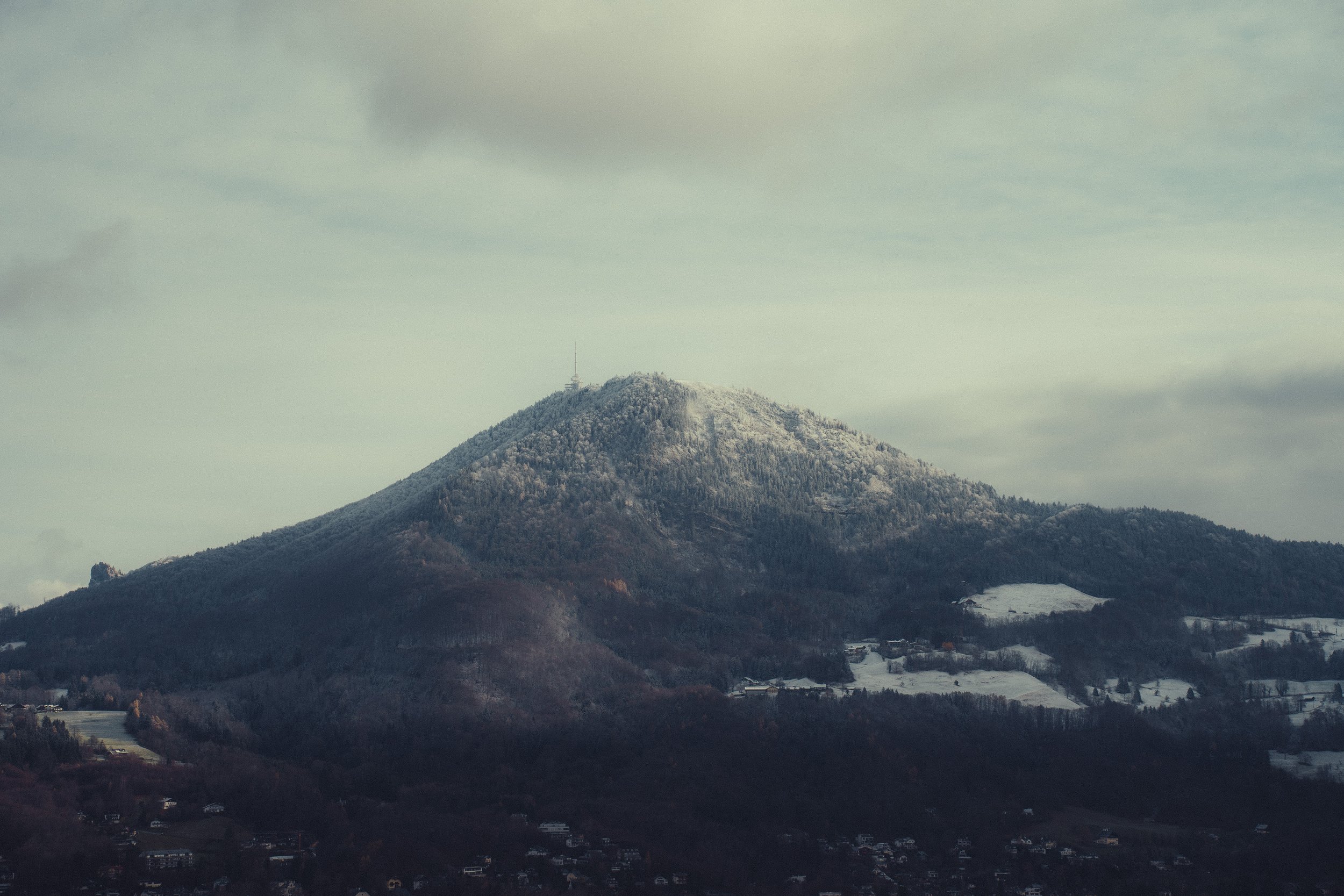 A snow-capped mountain with a forested slope and a small town at the base, under a cloudy sky.