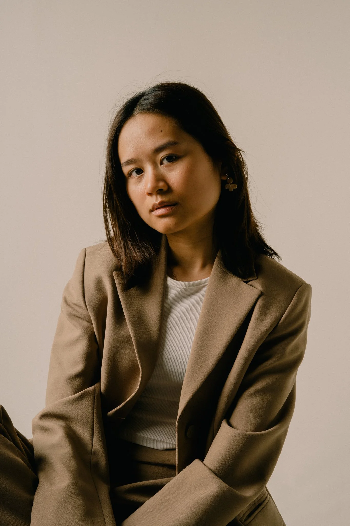 A young woman with shoulder-length dark hair wearing a tan blazer and earrings, posing against a plain beige background.