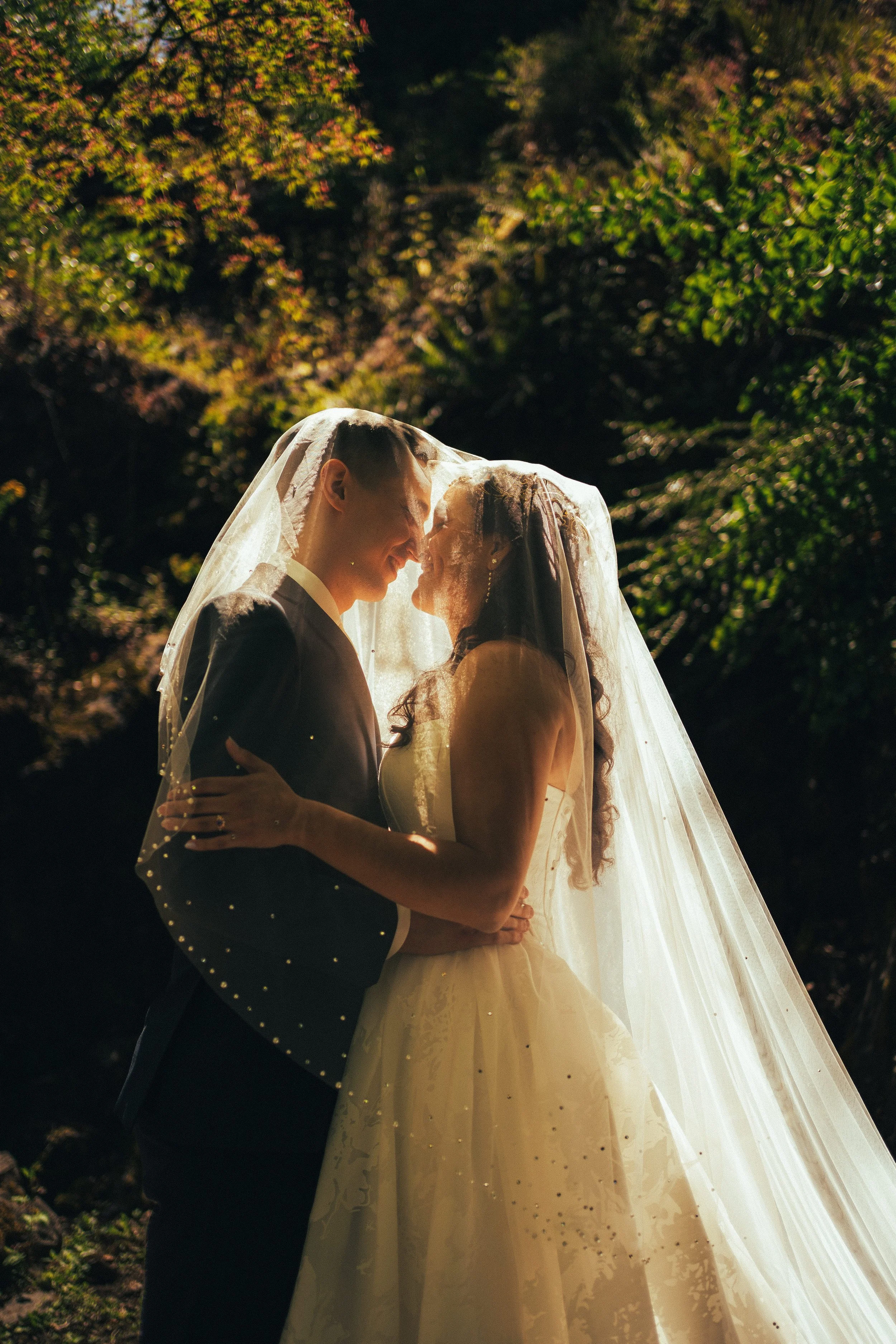 A bride and groom embrace under a long veil, standing outdoors with trees in the background, during sunset.