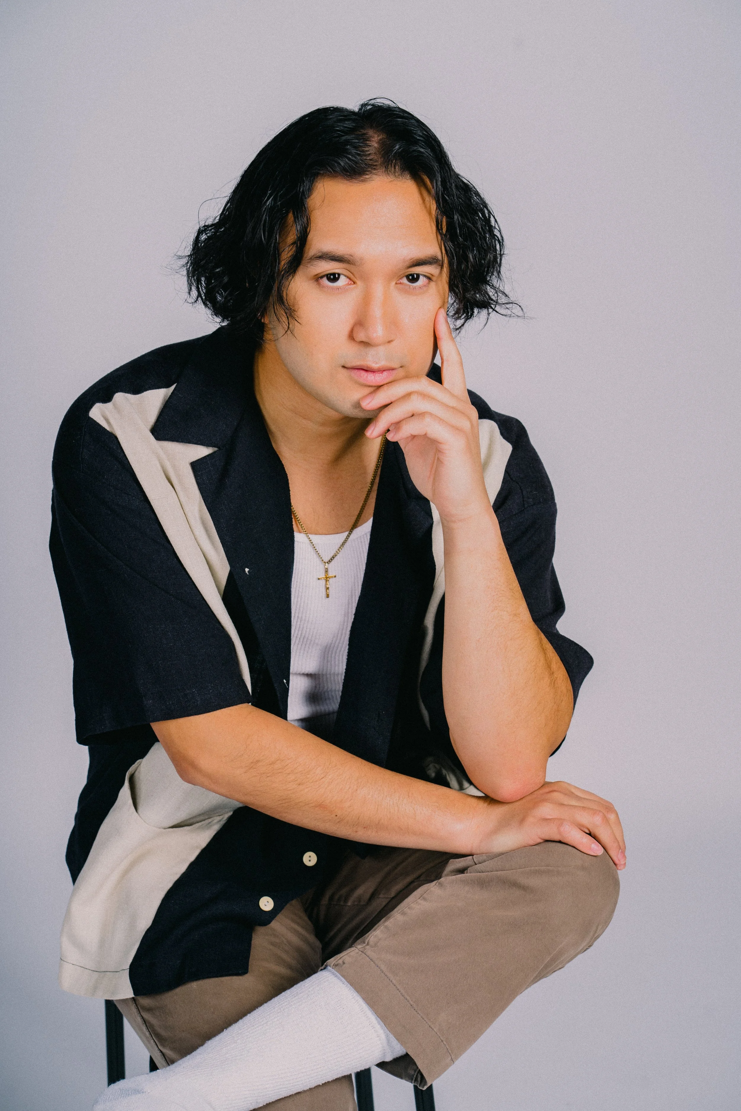 A man with black curly hair and light skin, sitting on a stool, wearing a black and beige button-up shirt, white undershirt, light brown pants, white socks, and a gold cross necklace, resting his chin on his hand and looking into the camera.