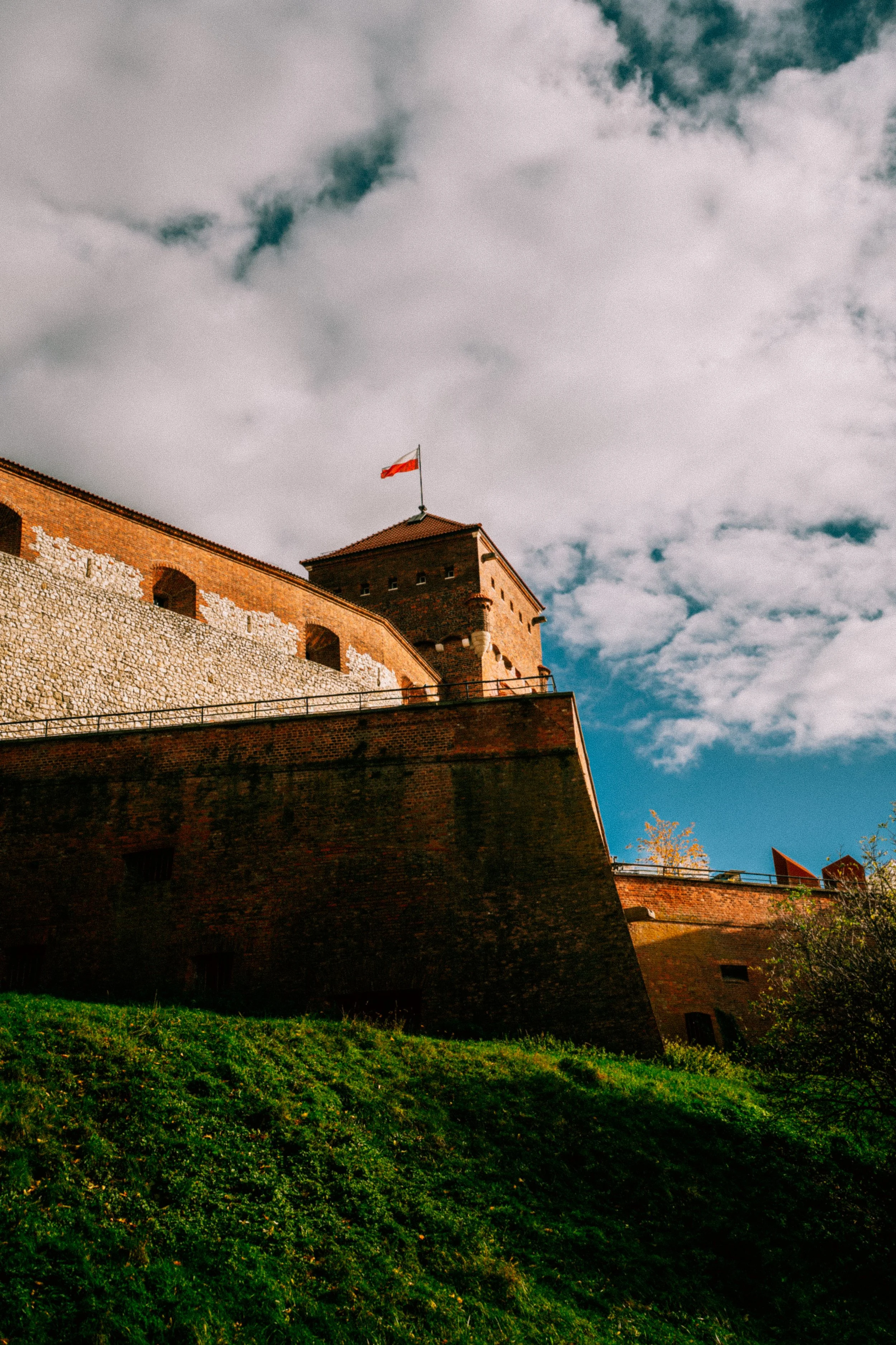 A historic brick fortress with a Polish flag flying on the top, under a partly cloudy sky, with green grass in the foreground.