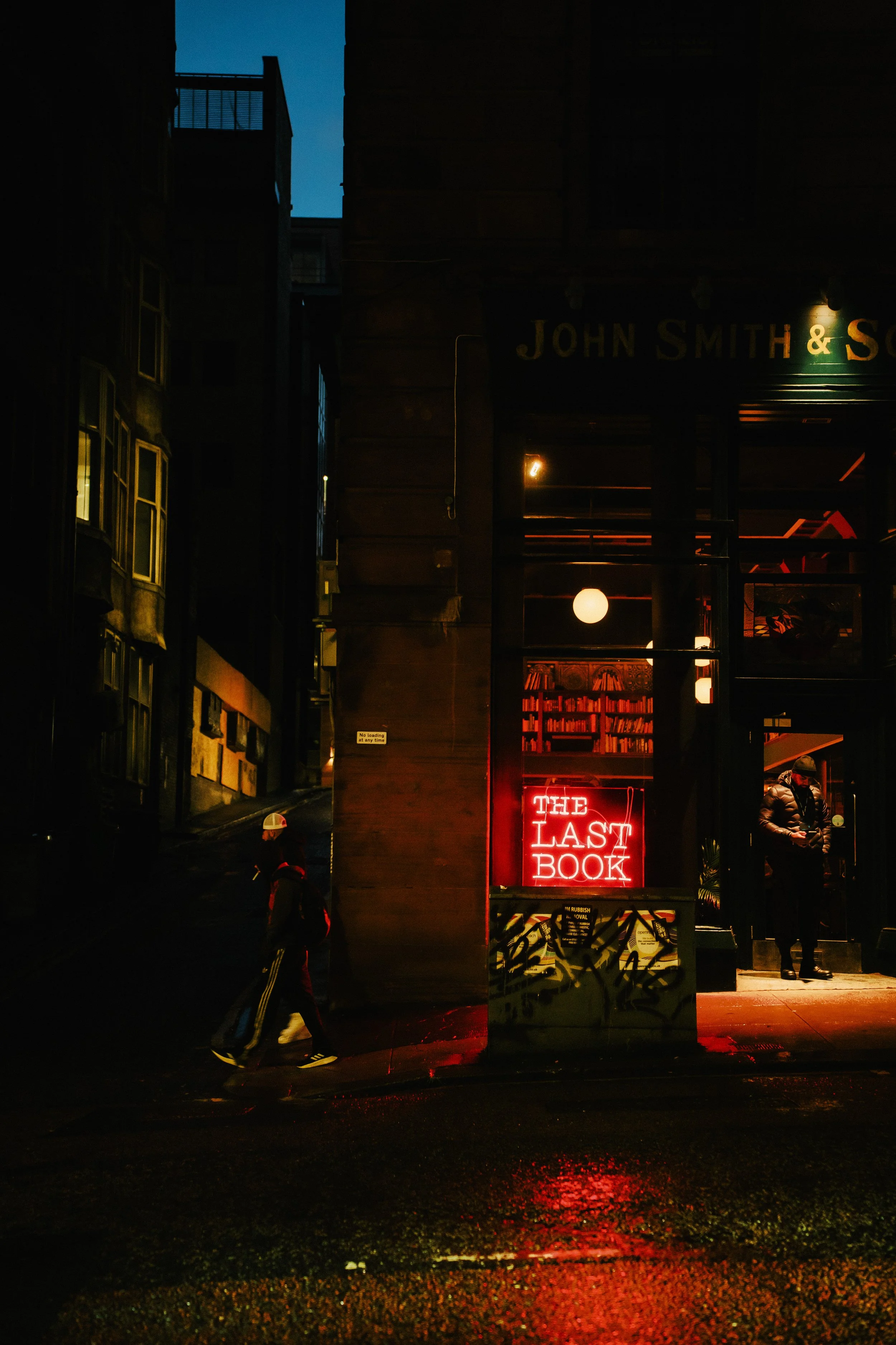 Nighttime street scene featuring a bookstore with a neon sign reading "The Last Book". A person walks on the sidewalk left, and a few people stand outside the bookstore entrance on the right. The store interior has warm lighting and bookshelves visib