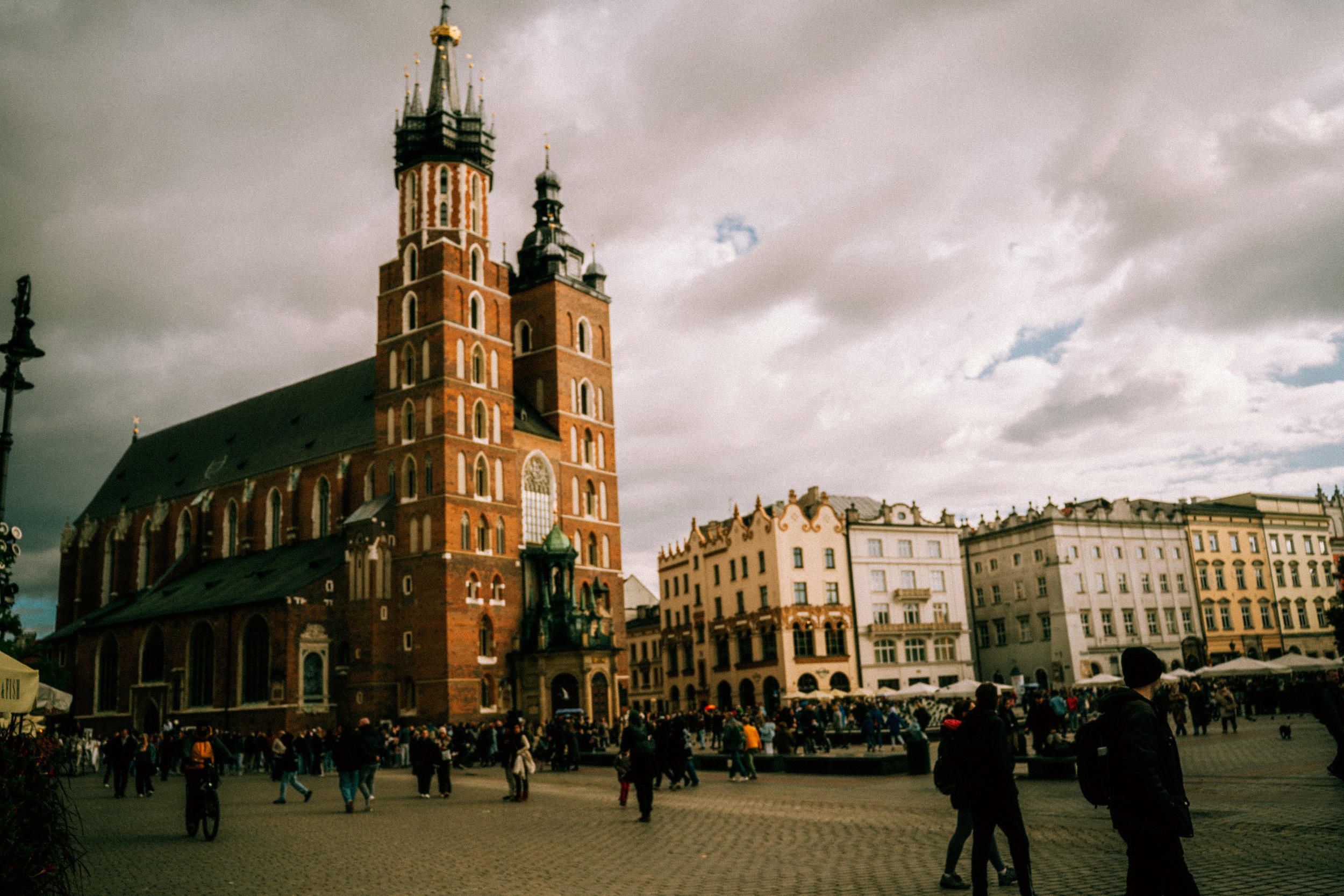 A large brick church with tall steeples in a busy city square, surrounded by historic buildings and many pedestrians.