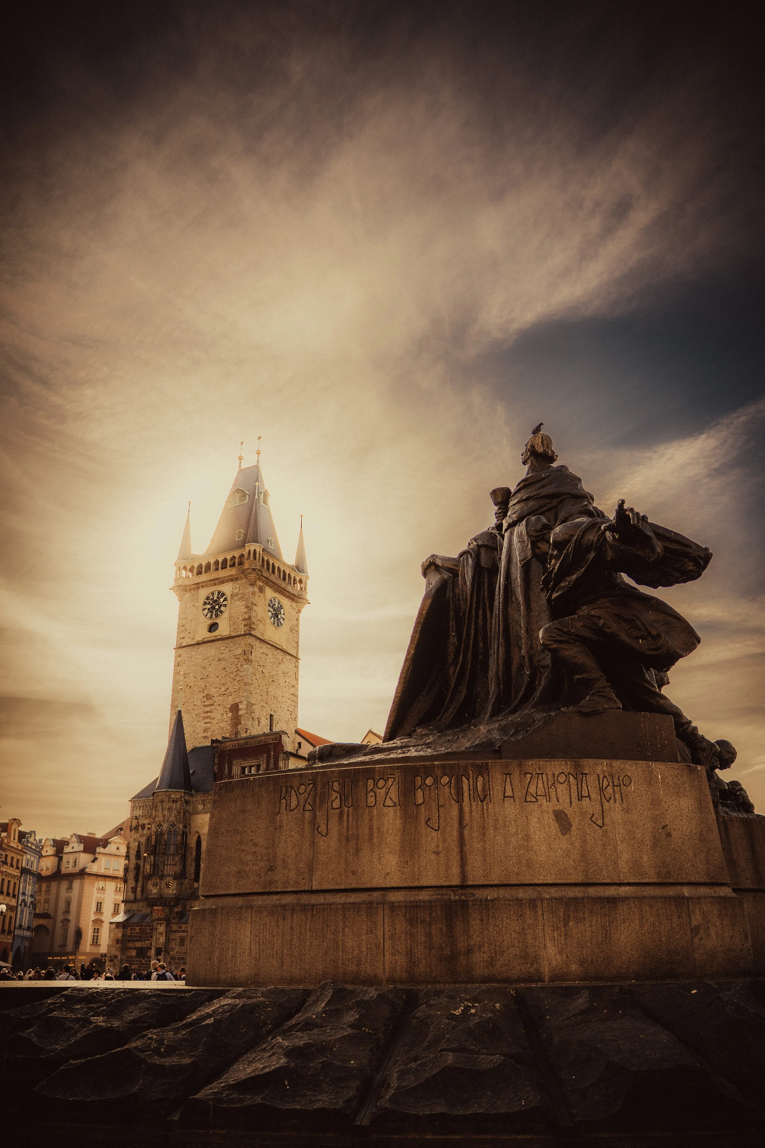 Historic clock tower with pointed spires and a statue of a knight on horseback in the foreground, in a European city square during sunset.