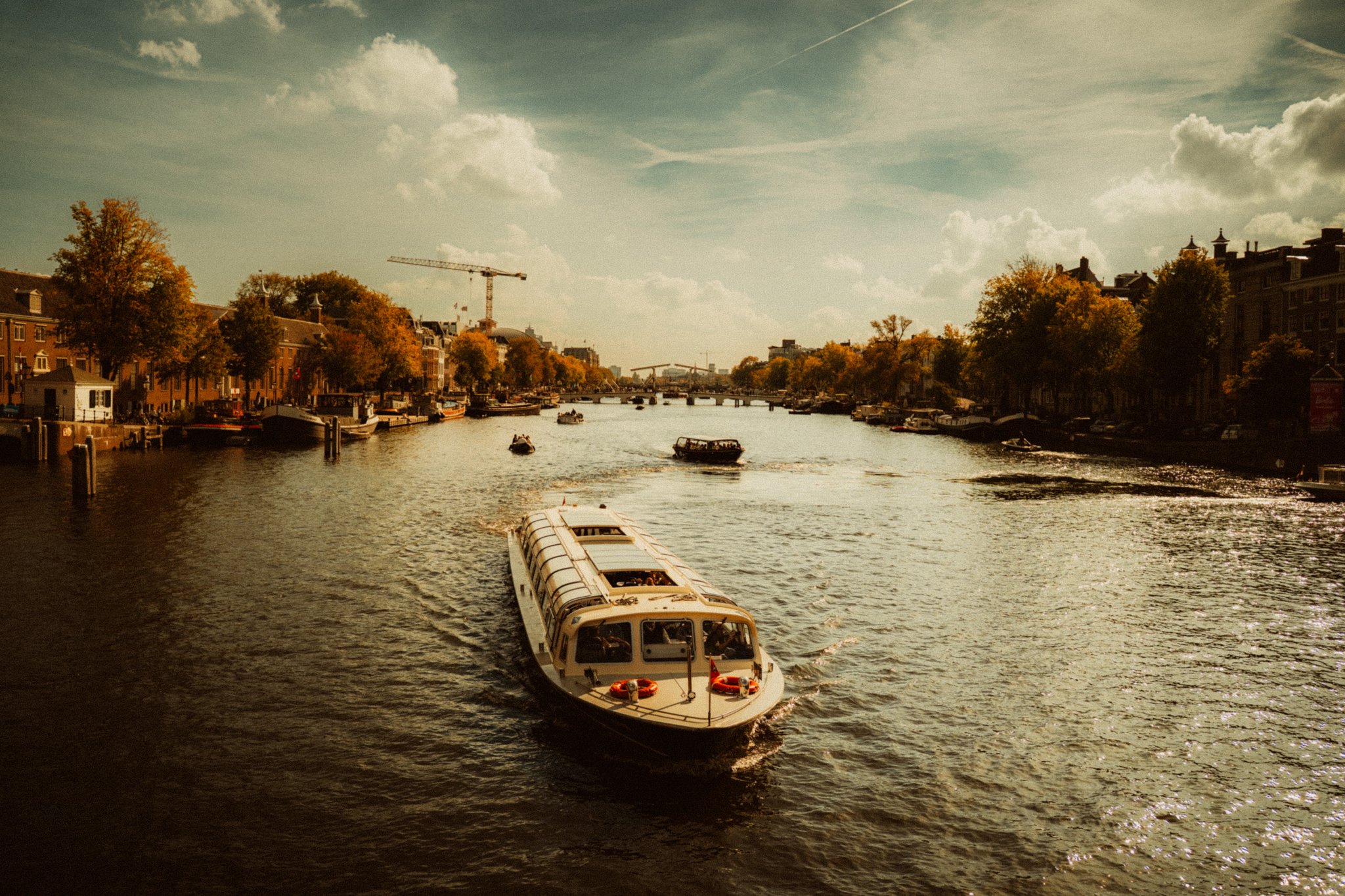 A river scene in Amsterdam, Netherlands ,during daytime with boats, trees, and buildings on both sides. The sky is partly cloudy with a warm, golden hue.