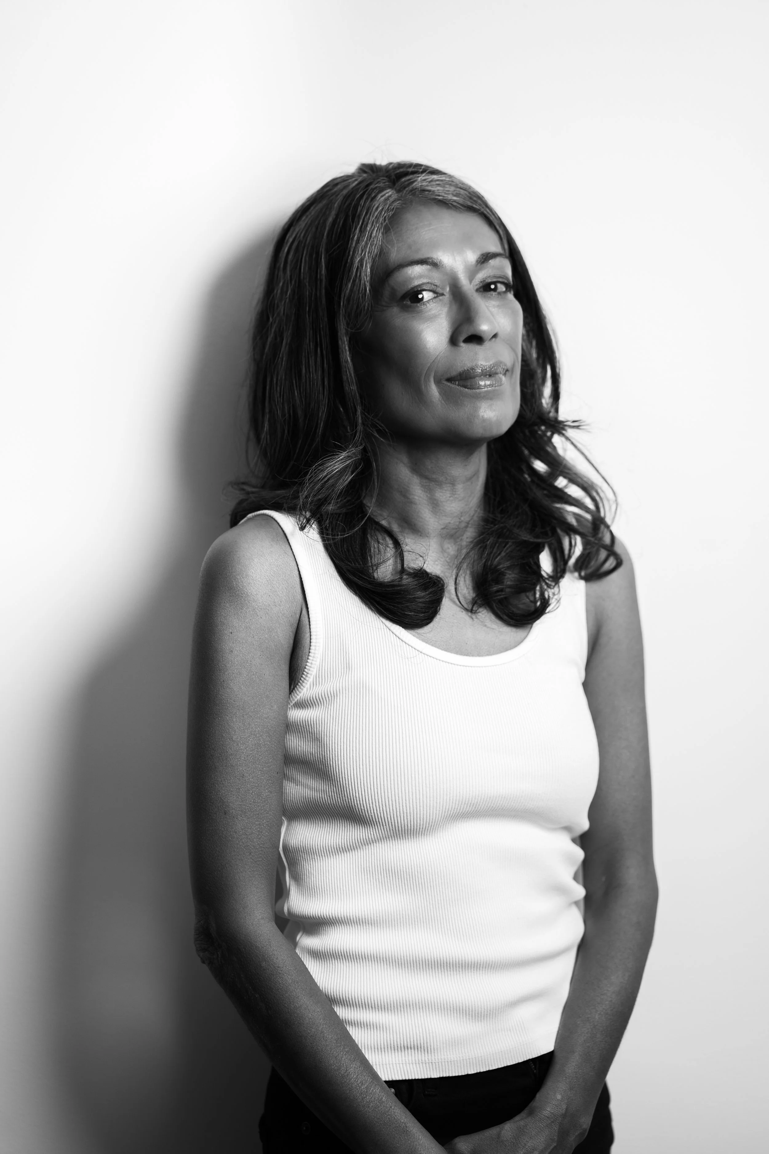 Black and white portrait of a woman with shoulder-length wavy hair, wearing a sleeveless white top, standing against a plain light background, looking confidently at the camera.