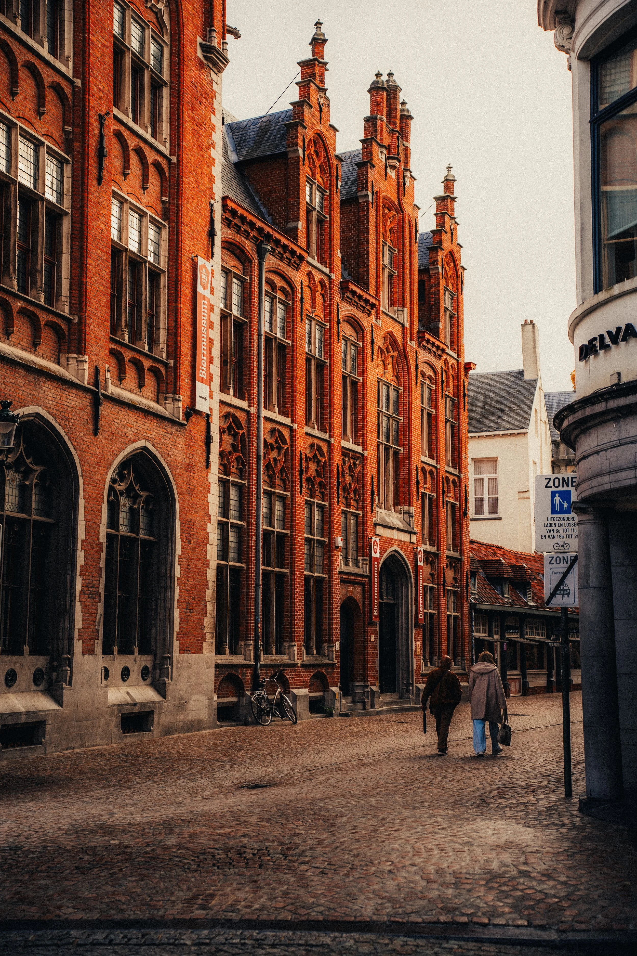 A street scene featuring a red brick Gothic-style building with large windows and decorative architectural details. Two people are walking on the cobblestone street, and there is a bicycle leaning against the building. Street signs are visible on the