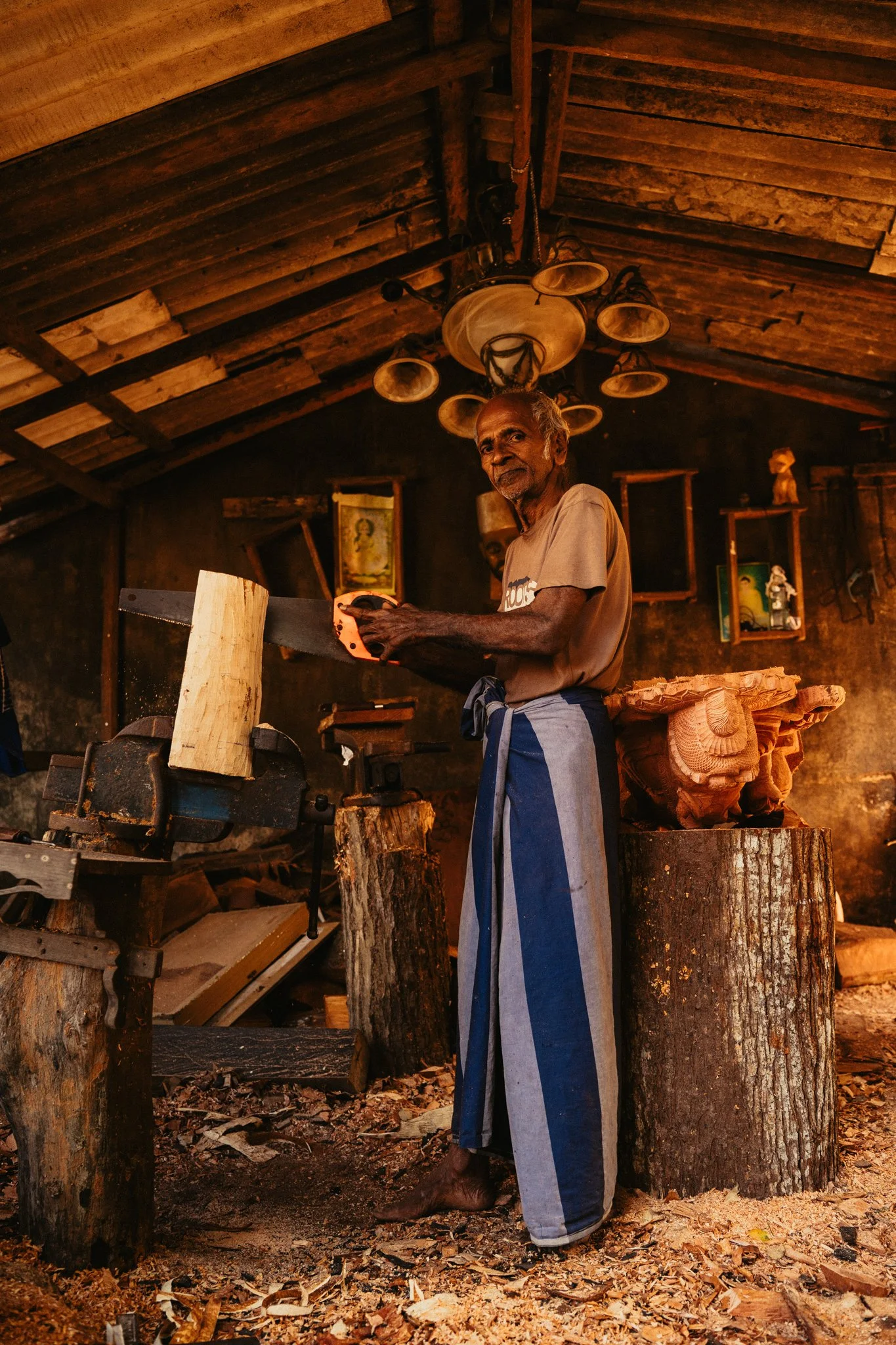 An elderly man with gray hair and a beard, wearing a beige t-shirt and blue-striped sarong, is sawing a piece of wood in a rustic woodworking shop. The shop has a wooden ceiling with hanging lights and shelves with small objects and pictures on the d