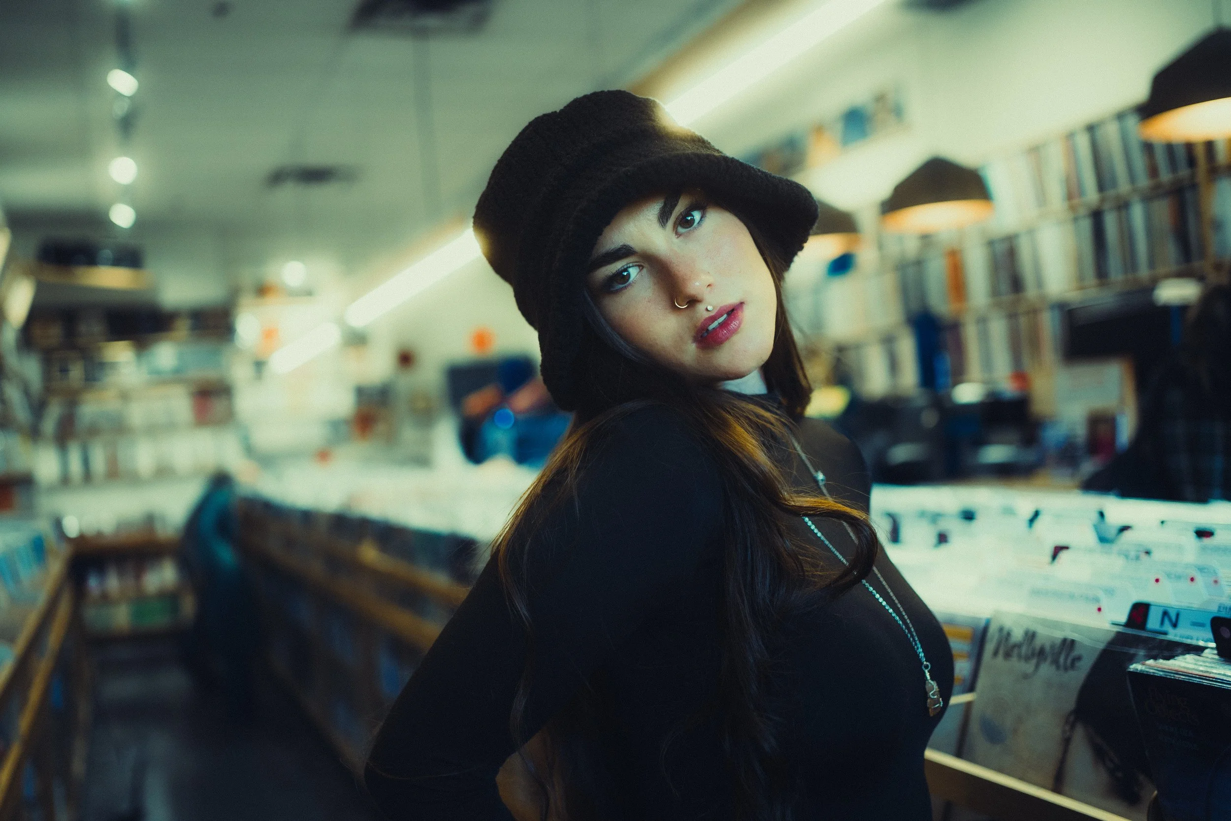 Young woman with dark hair, wearing a black hat and black top, looking at the camera inside record store shelves filled with vinyl records.