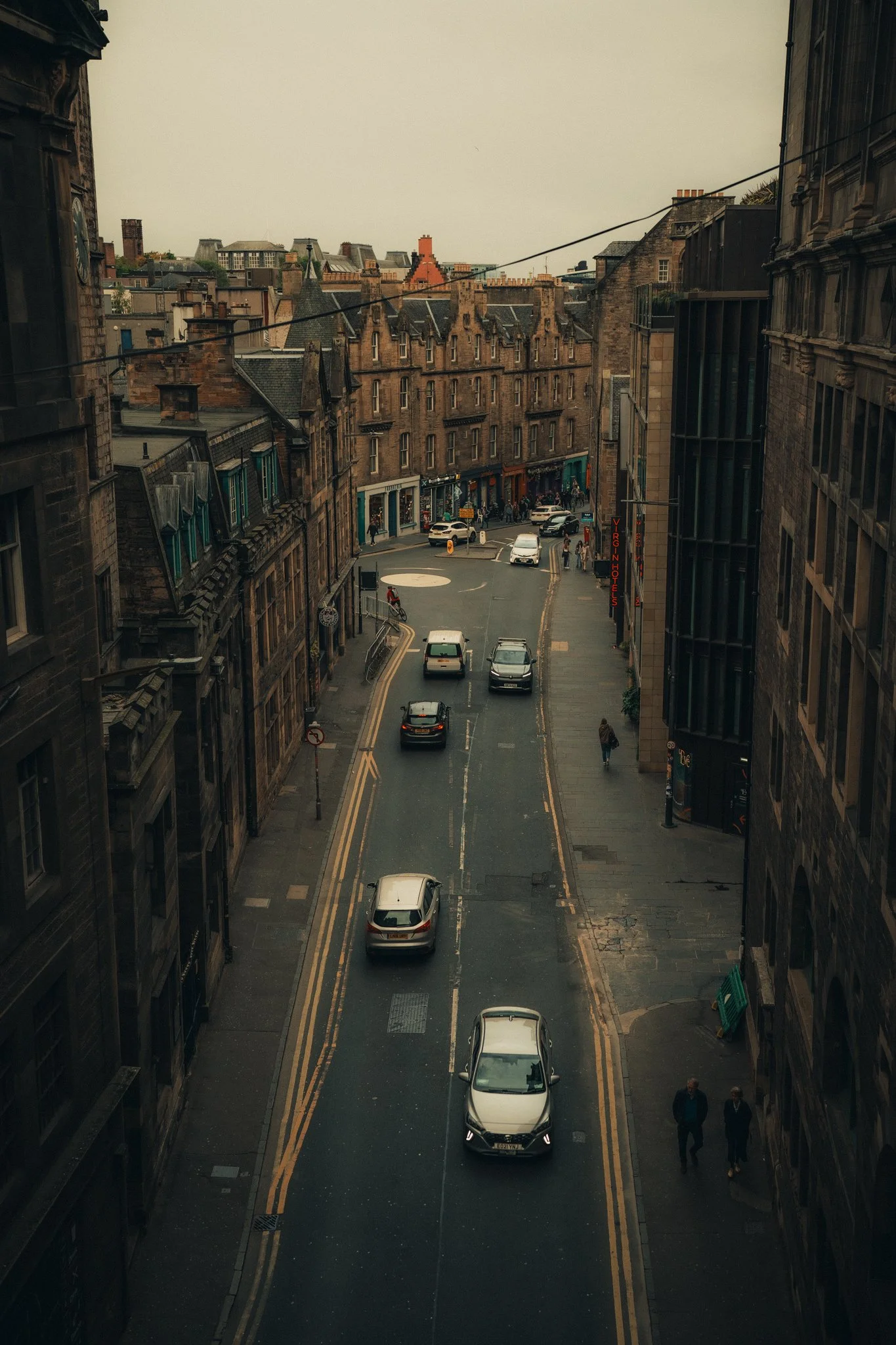 A narrow city street in Scotland with parked and moving cars, pedestrians walking along the sidewalks, and older brick buildings on both sides.