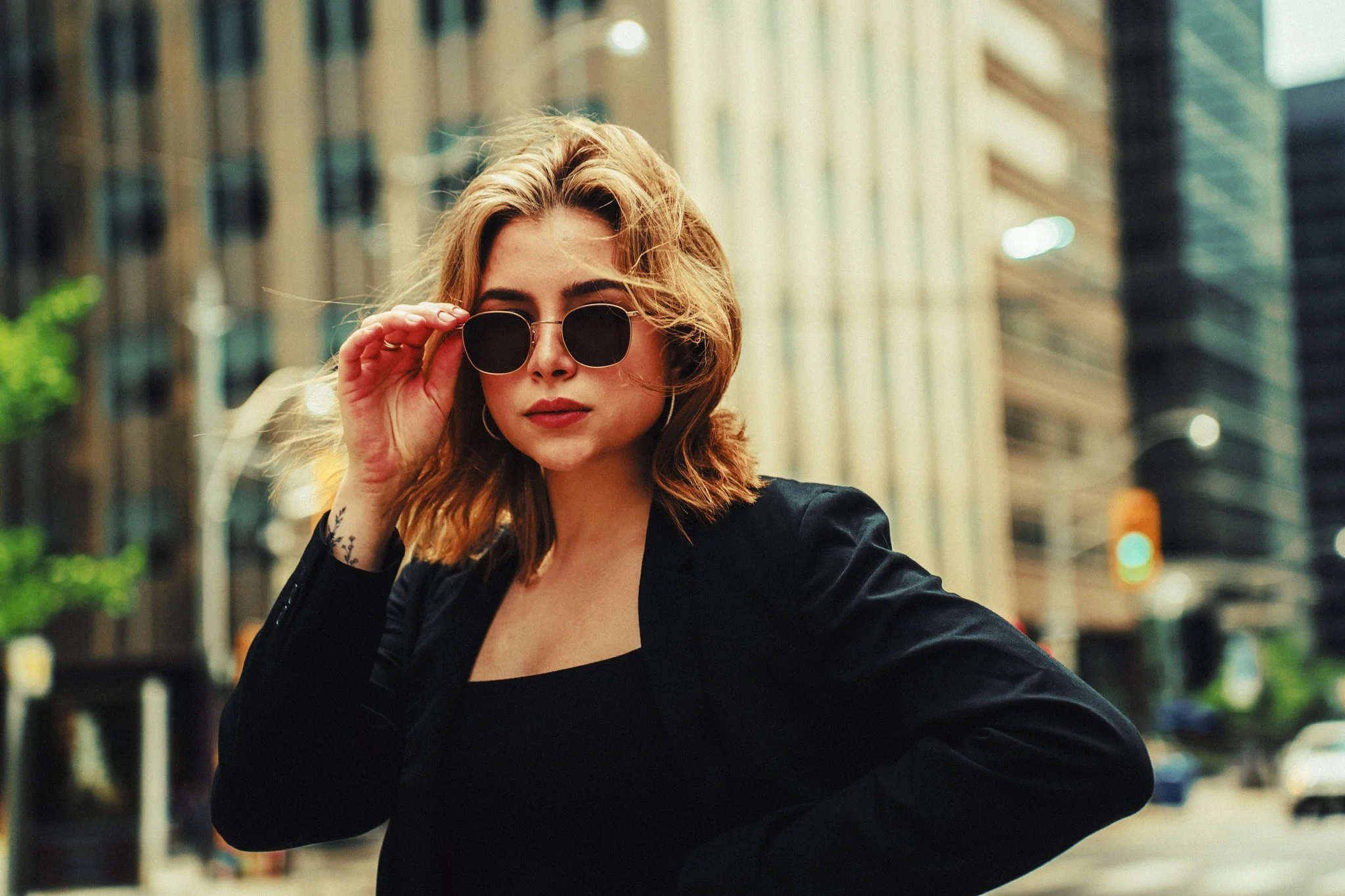 A woman with blonde hair wearing a black blazer and black top, holding sunglasses in a city street scene, with tall buildings and blurred traffic in the background.