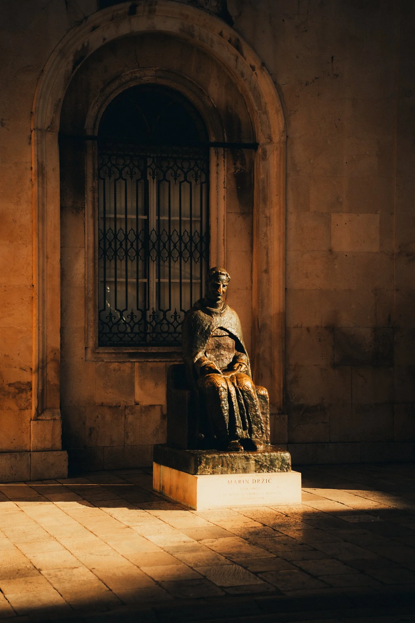 A bronze statue of a seated man with a cape, located outside a building with an arched window and iron bars. The statue is on a white pedestal with an inscription, and warm lighting highlights the figure against a stone wall.