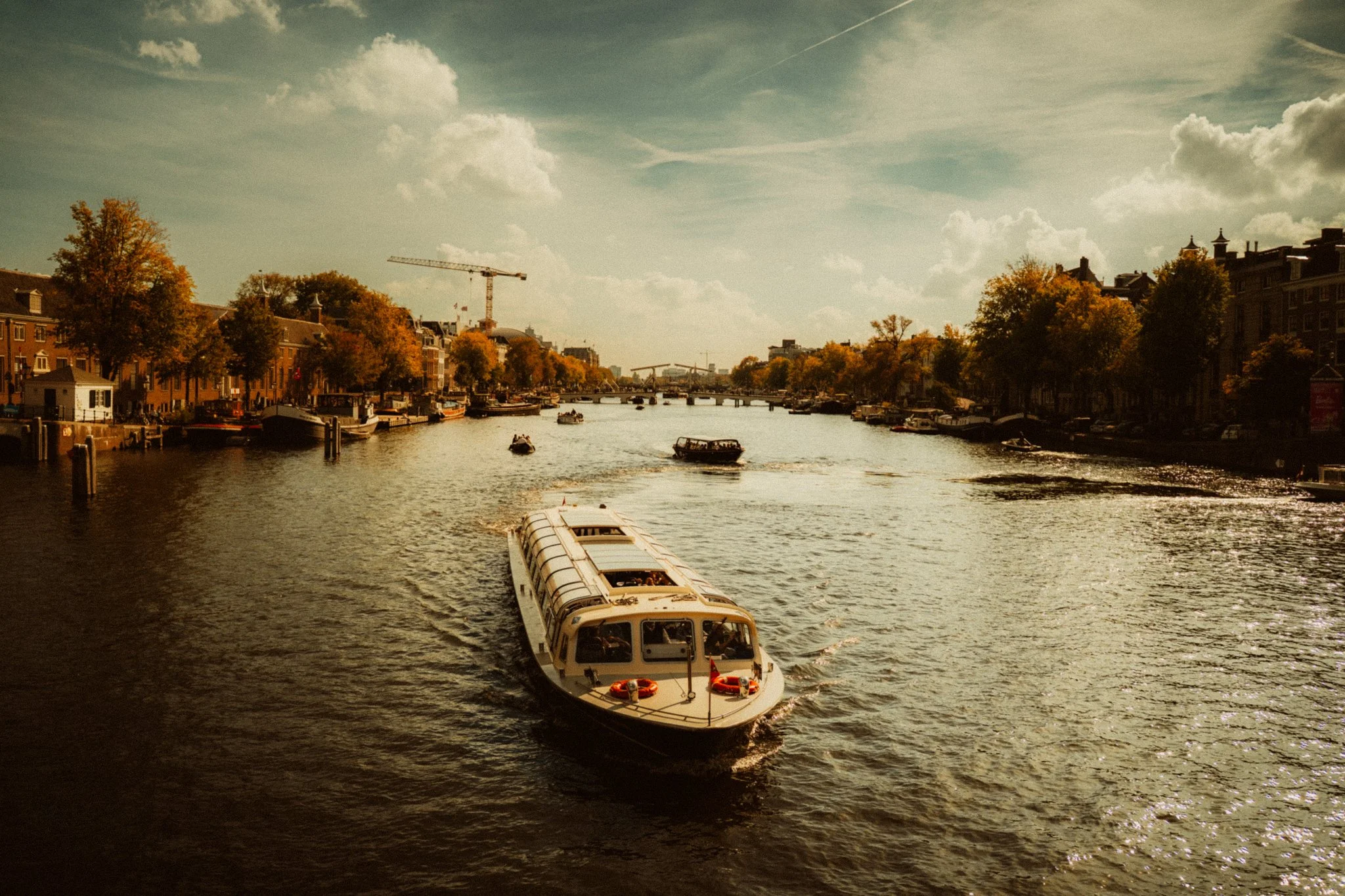 A canal scene with boats, trees with autumn foliage, and buildings lining the water under a partly cloudy sky.
