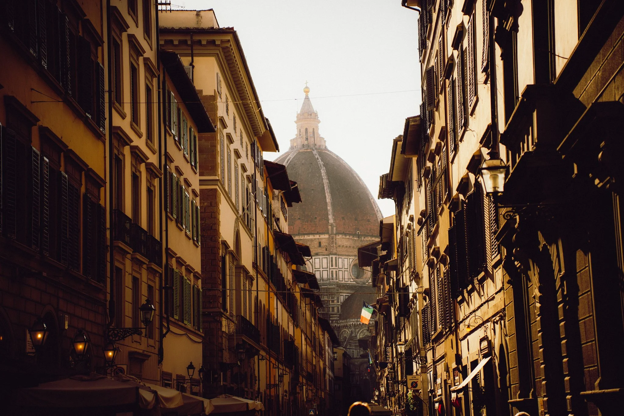 A narrow street lined with tall, historic buildings with dark shutters. The street leads to the Florence Cathedral's dome in the background.