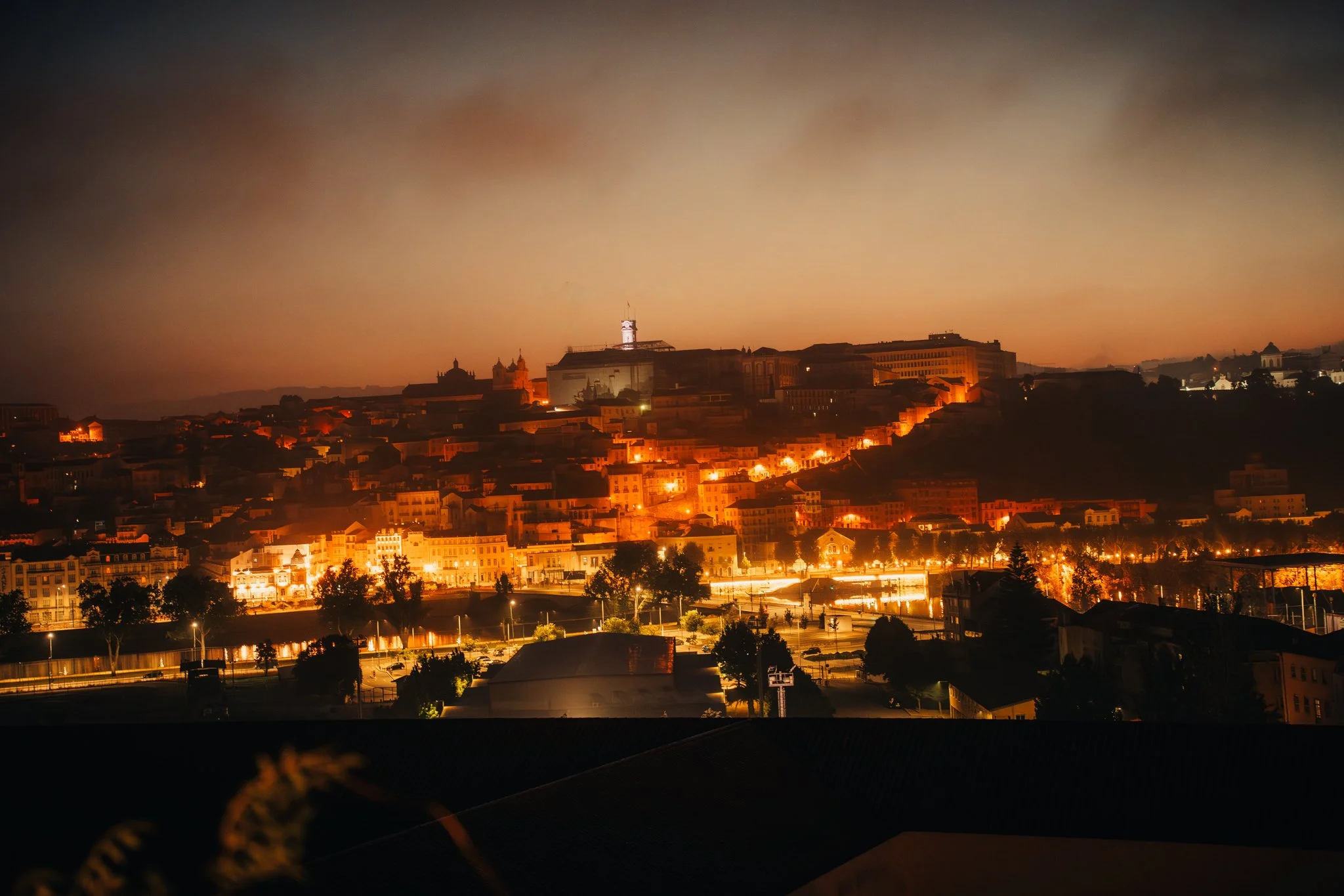 Nighttime cityscape with illuminated buildings and streets on a hillside, with a clear sky.