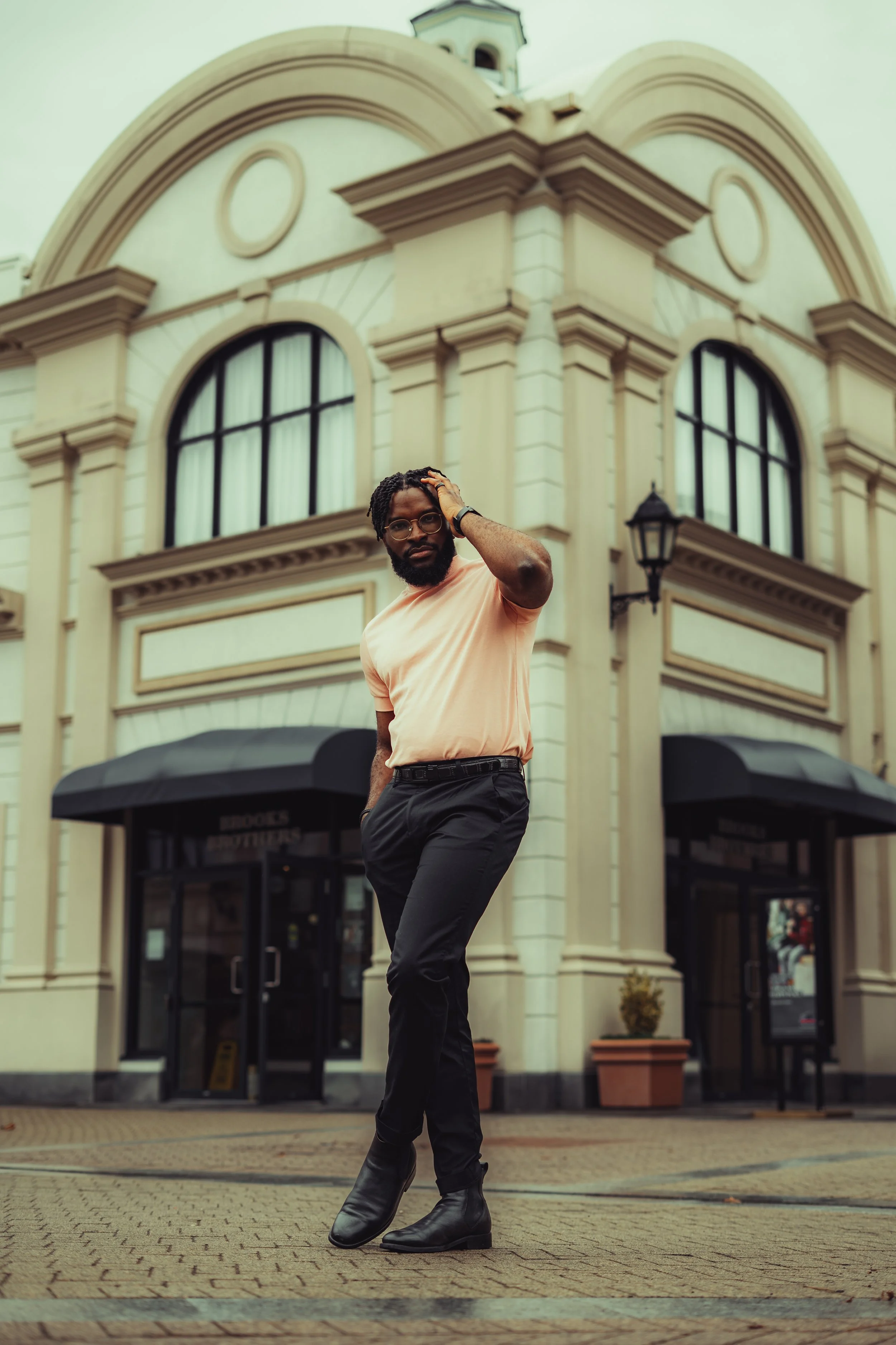 A man with glasses, a beard, and dreadlocks standing in front of a beige, ornate building with large windows and black awnings. He is wearing a peach-colored shirt and black pants with black boots, posing with one hand on his head.