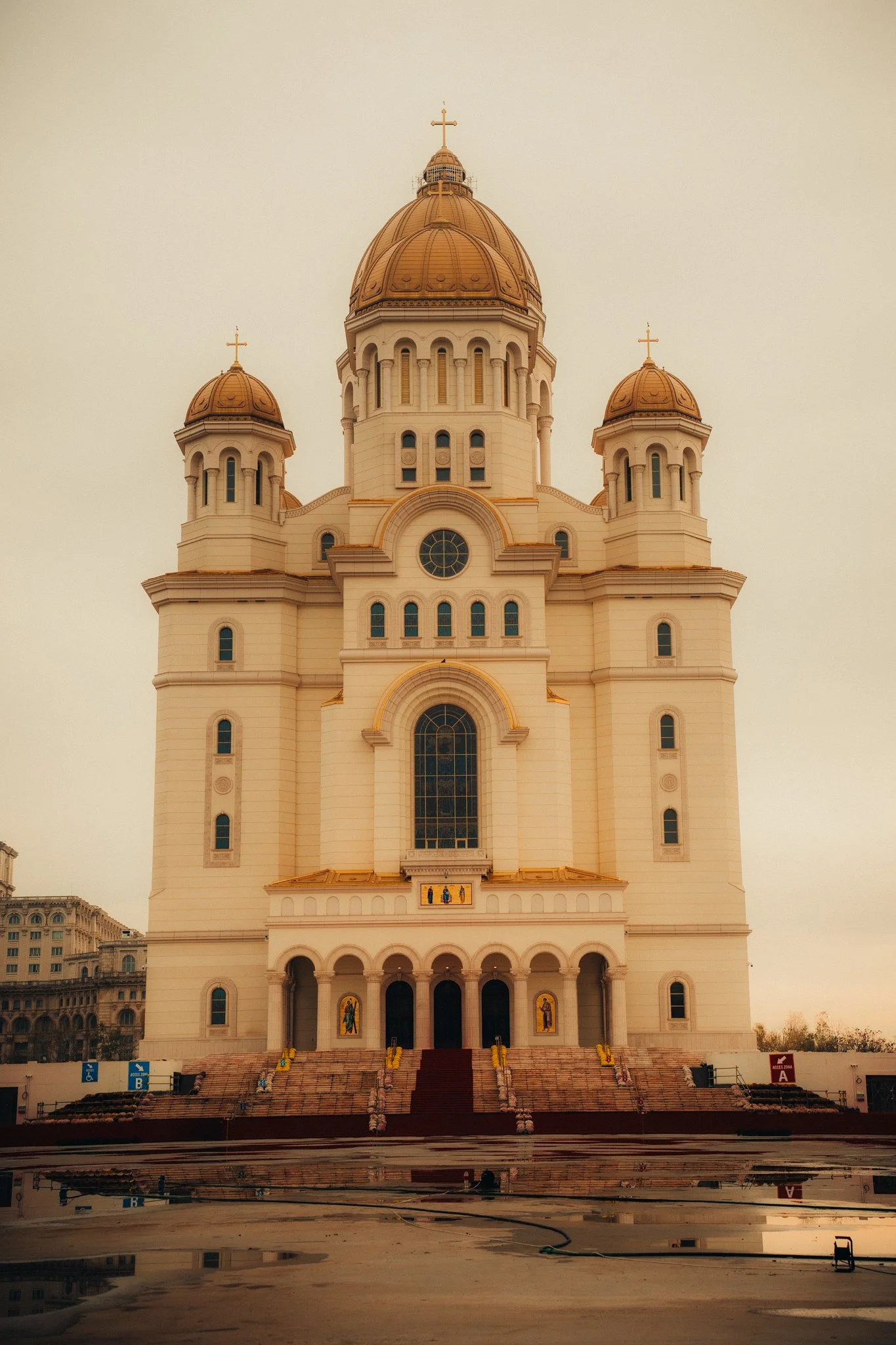 The image shows a large, ornate church with a central dome and four smaller domes, all with crosses on top. The church has a beige exterior with gold accents and multiple arched windows and doors. The steps leading up to the entrance are reflected in