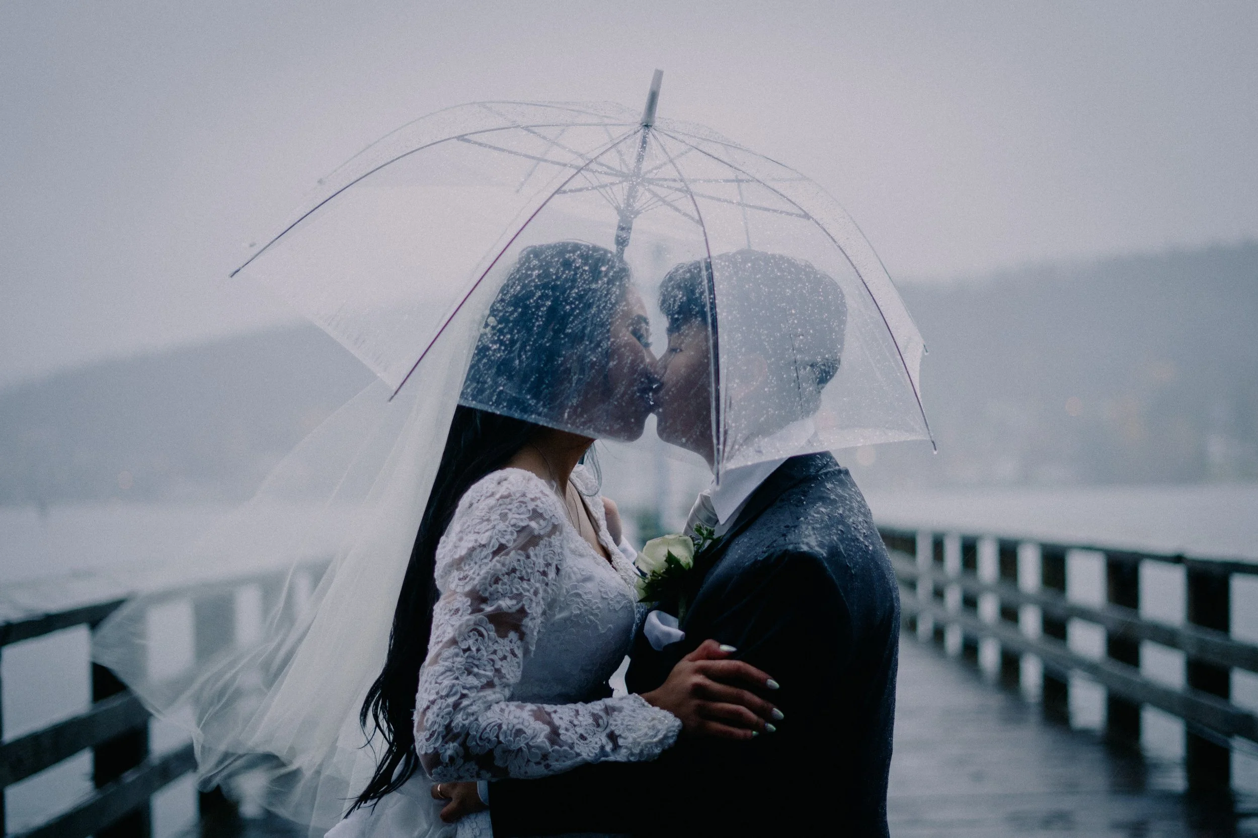 A bride and groom sharing a kiss under a transparent umbrella on a rainy day near a body of water, with a wooden pier in the background.