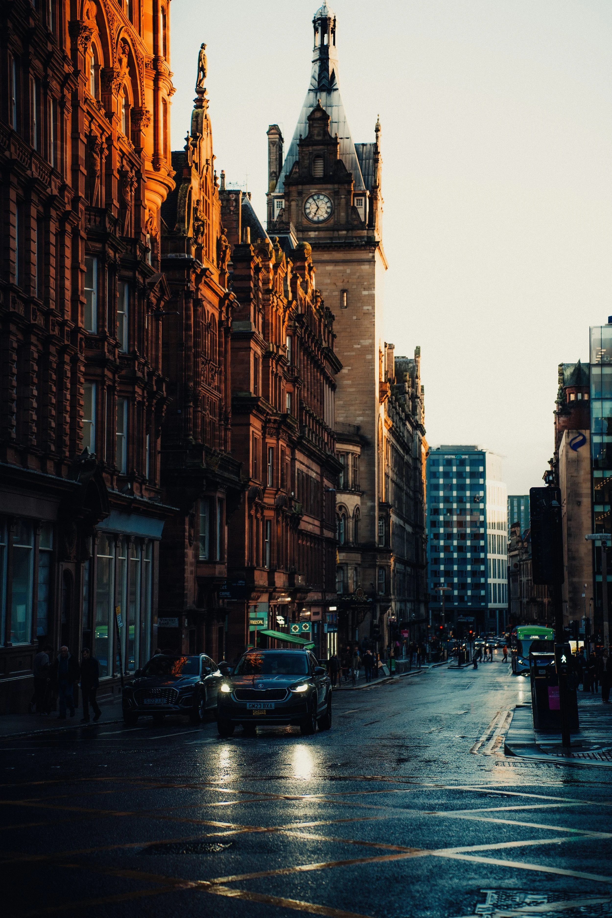 City street at sunset with historic buildings, cars, and pedestrians, wet pavement reflecting lights.