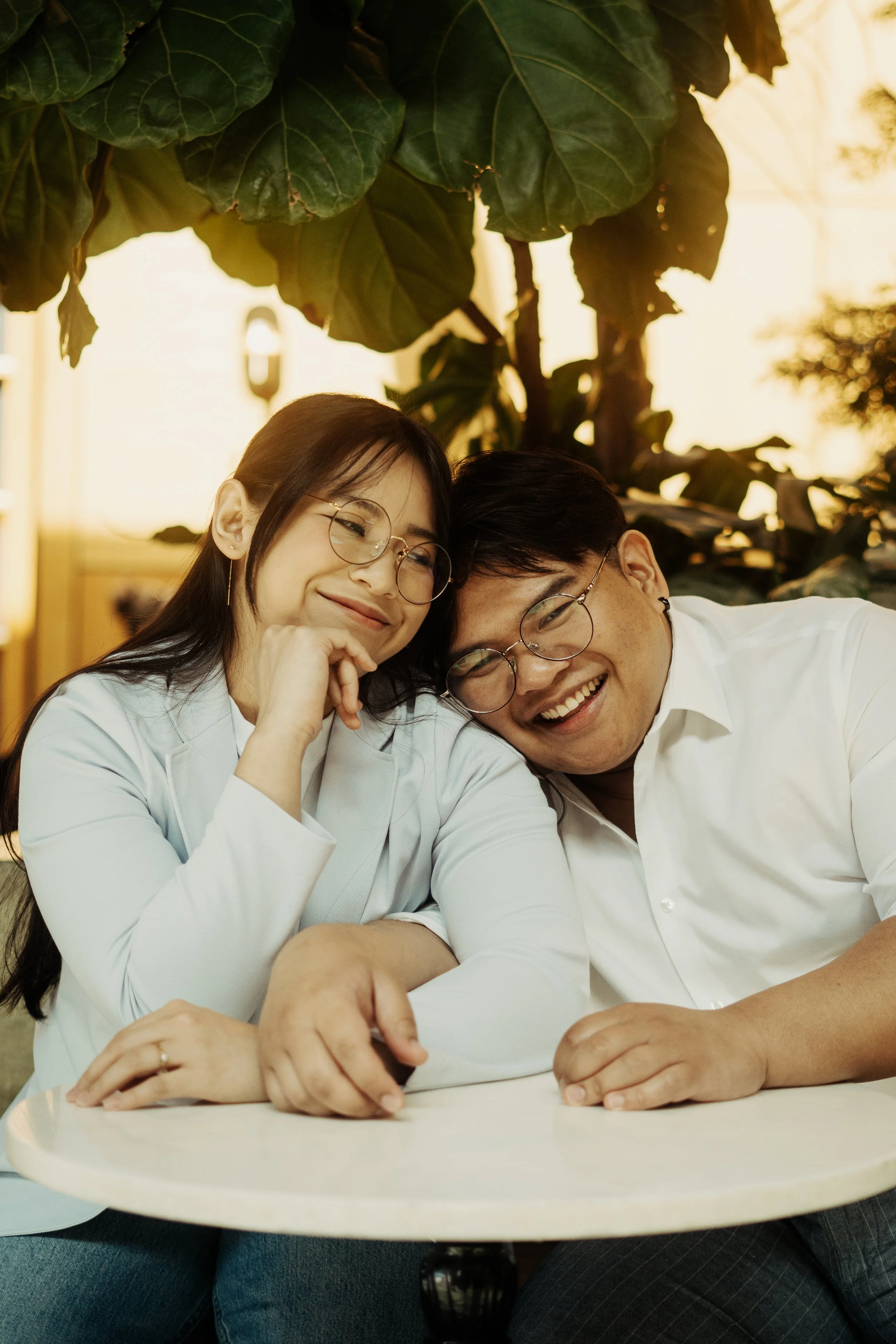 A young woman and a young man sit close together at a white table, smiling happily, with large green leaves overhead in a warmly lit indoor setting.