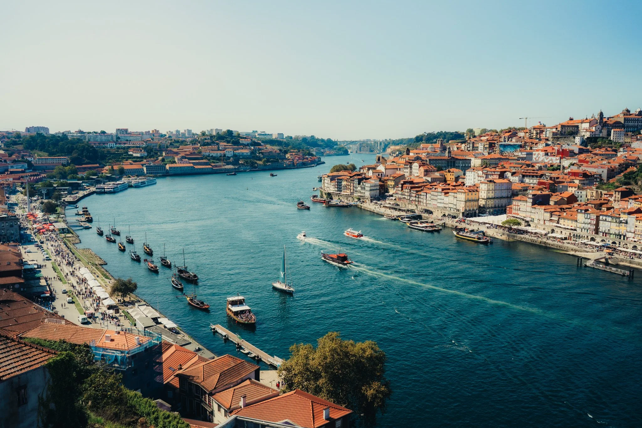 Porto, Portugal - A river with boats, surrounded by city buildings with terracotta roofs and trees, on a clear sunny day.