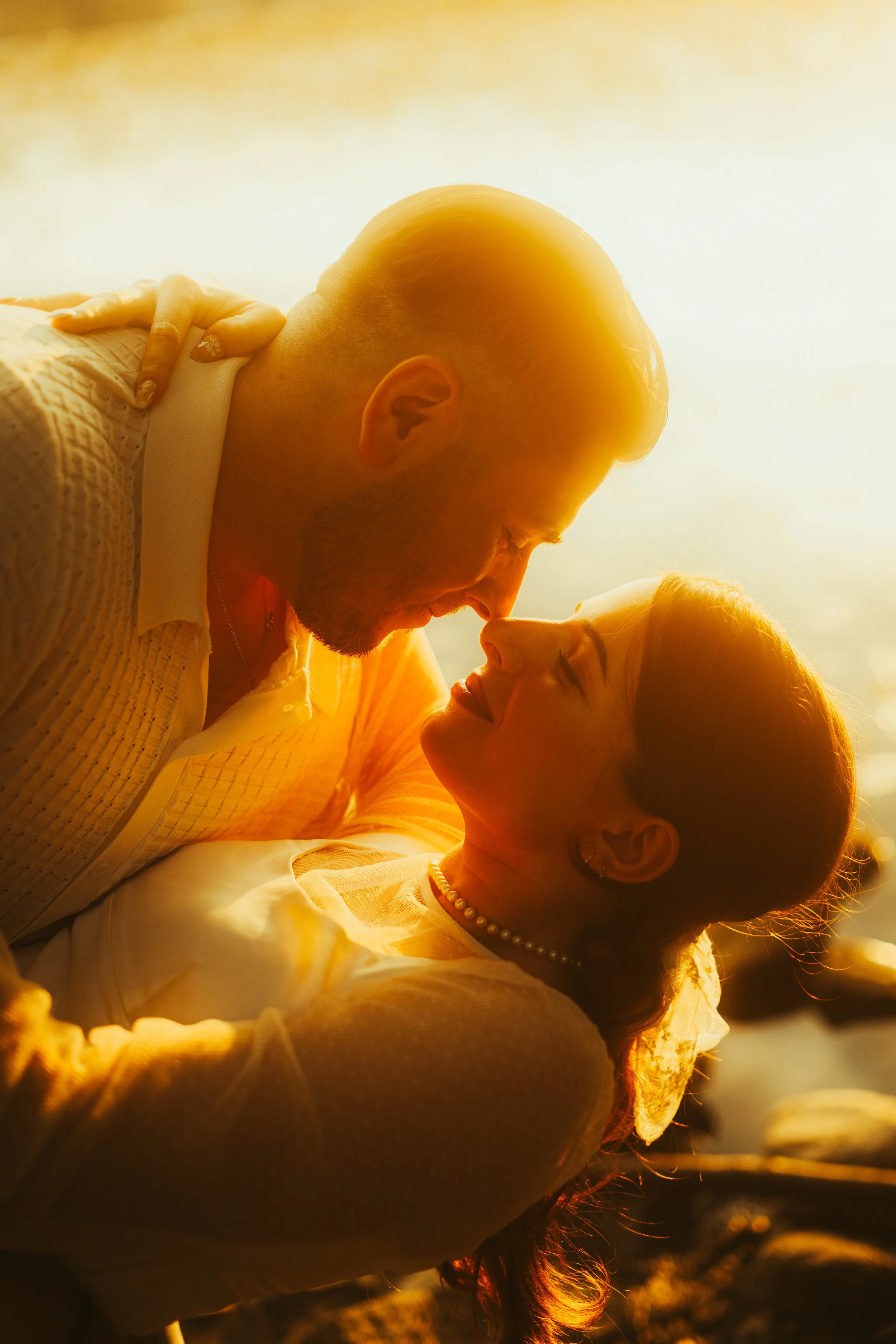 A couple close together, indoors, with sunlight behind them, nose to nose, smiling at each other.