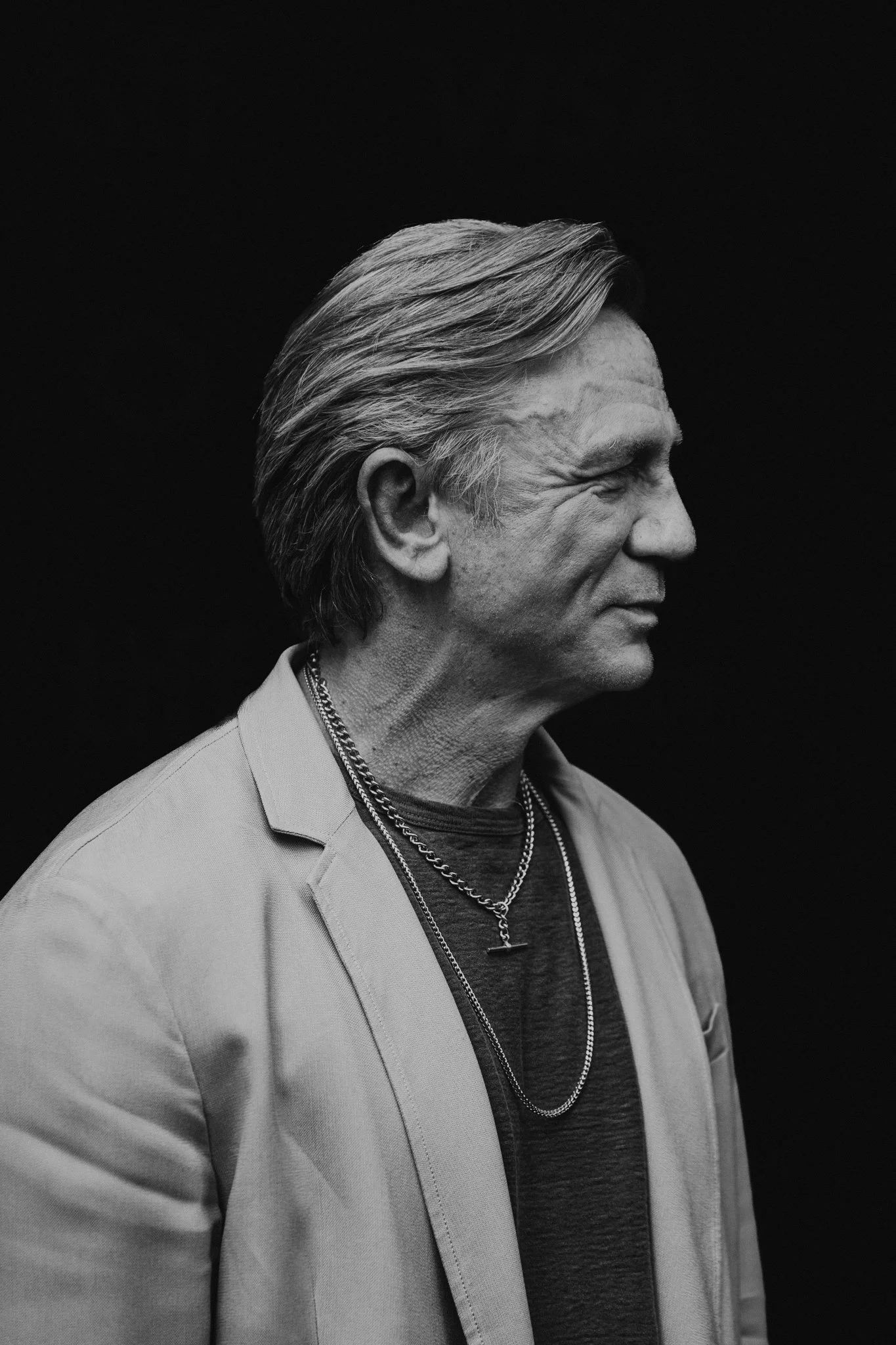 Black and white profile photo of Daniel Craig with slicked-back hair, wearing a light blazer and layered necklaces, against a black background.