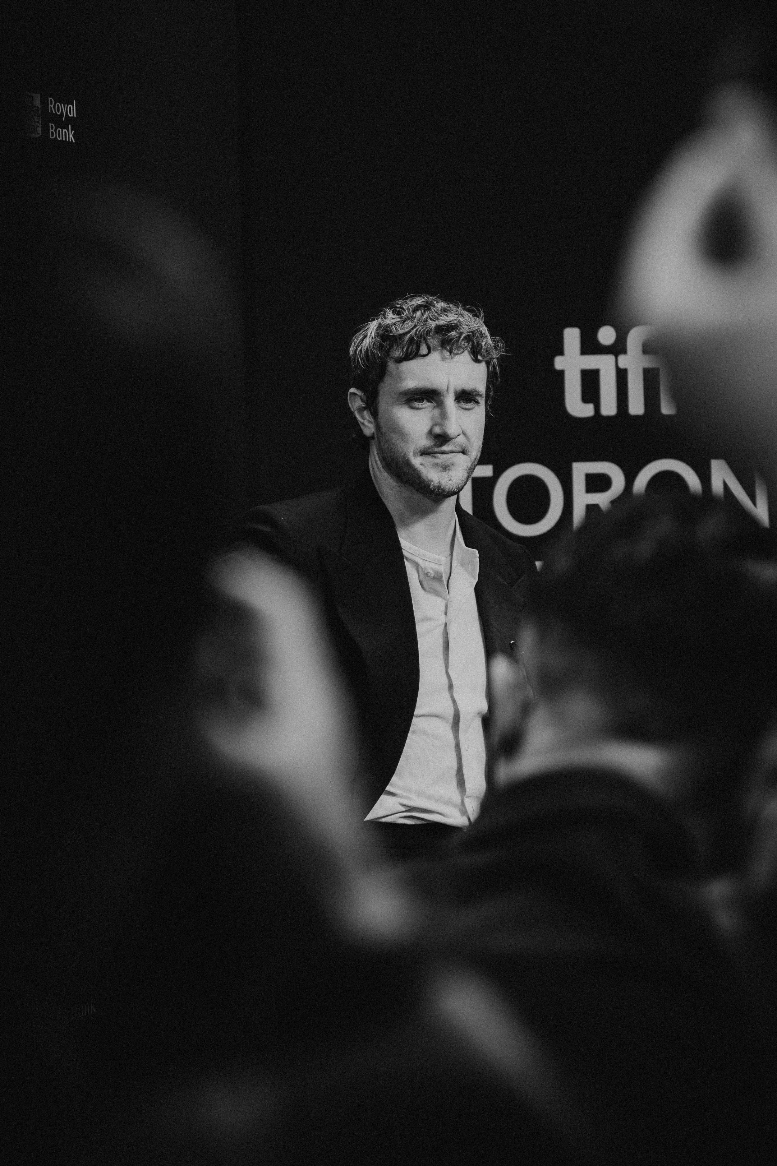 A black-and-white photograph of Paul Mescal with curly hair and a beard, wearing a suit jacket and a light-colored shirt, standing at a public event with a dark background displaying the words "tiff" and "TORON".