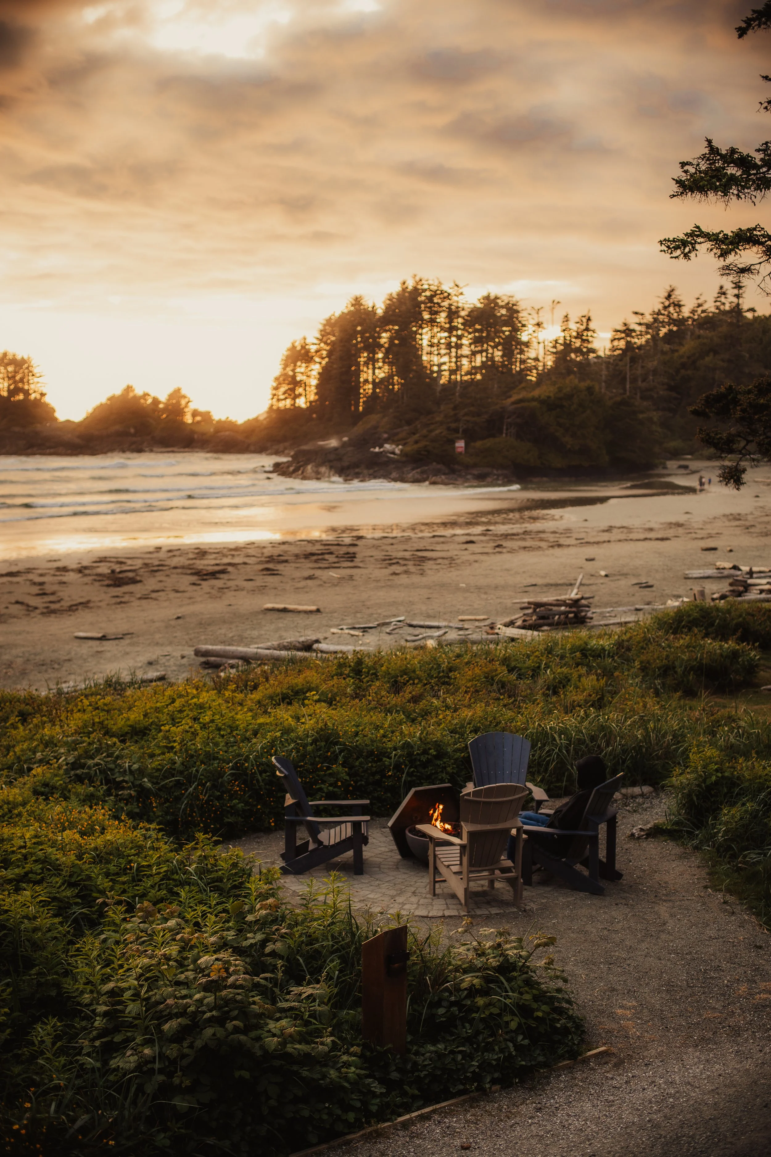 A beach at sunset with calm waves and a wooded coastline in the background. In the foreground, there is a small fire pit surrounded by four chairs on a gravel area, with some greenery and bushes around.