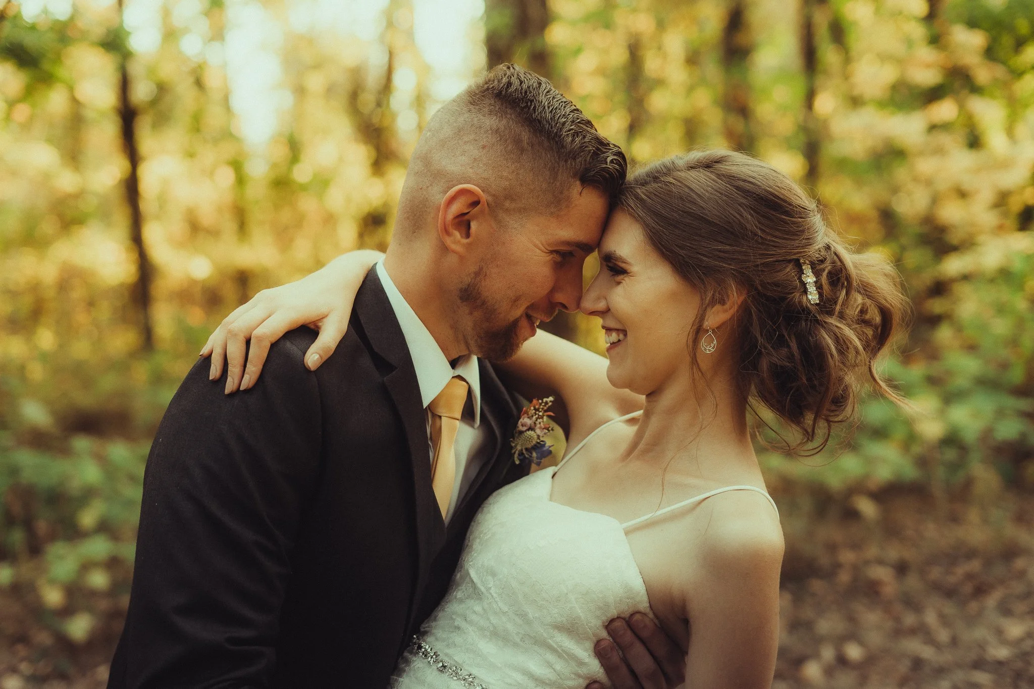 A newlywed couple embracing in a forest with fall foliage, smiling and touching foreheads.