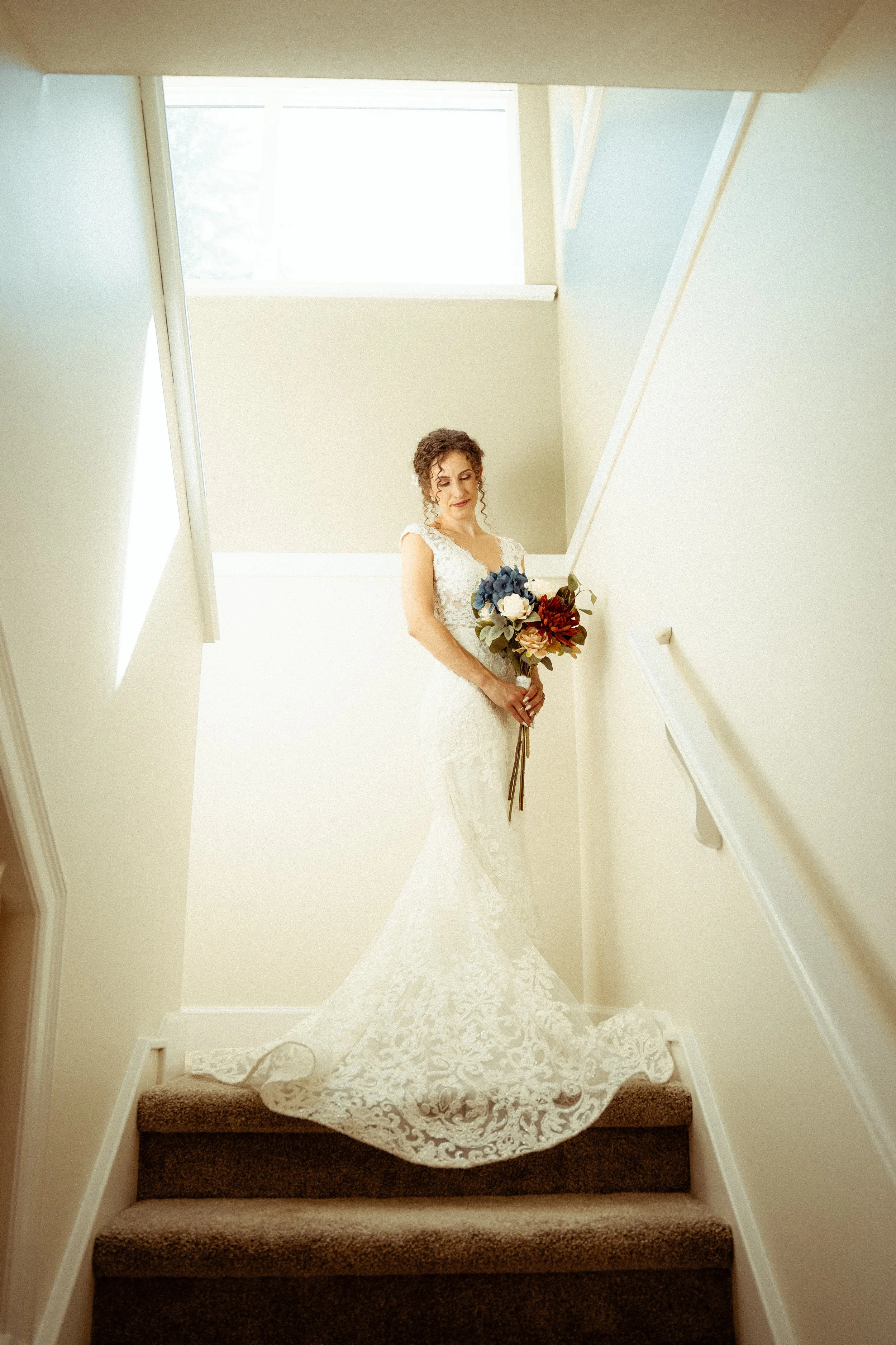 A bride in a white lace wedding dress holding a bouquet of flowers on a staircase.