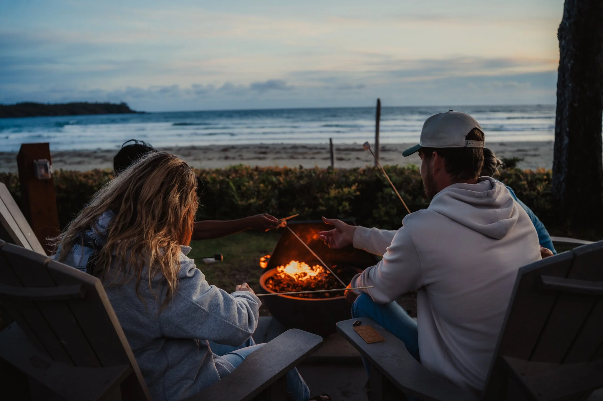 Group of people sitting around a campfire on the beach at sunset, roasting marshmallows.