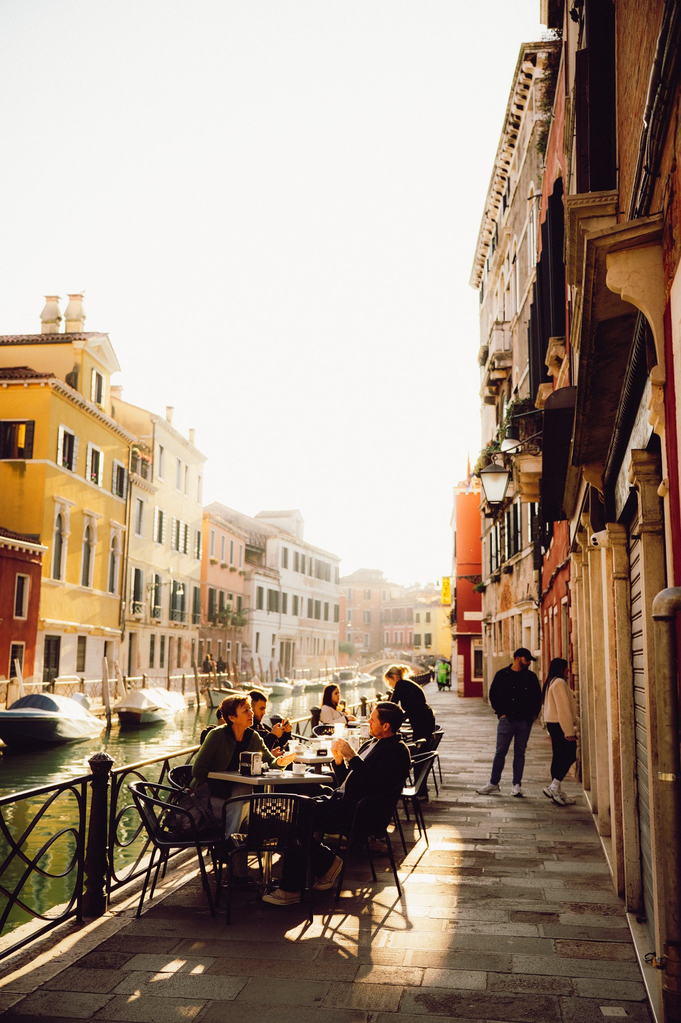 People dining outside along a canal in a European city at sunset, with colorful buildings and boats in the water.