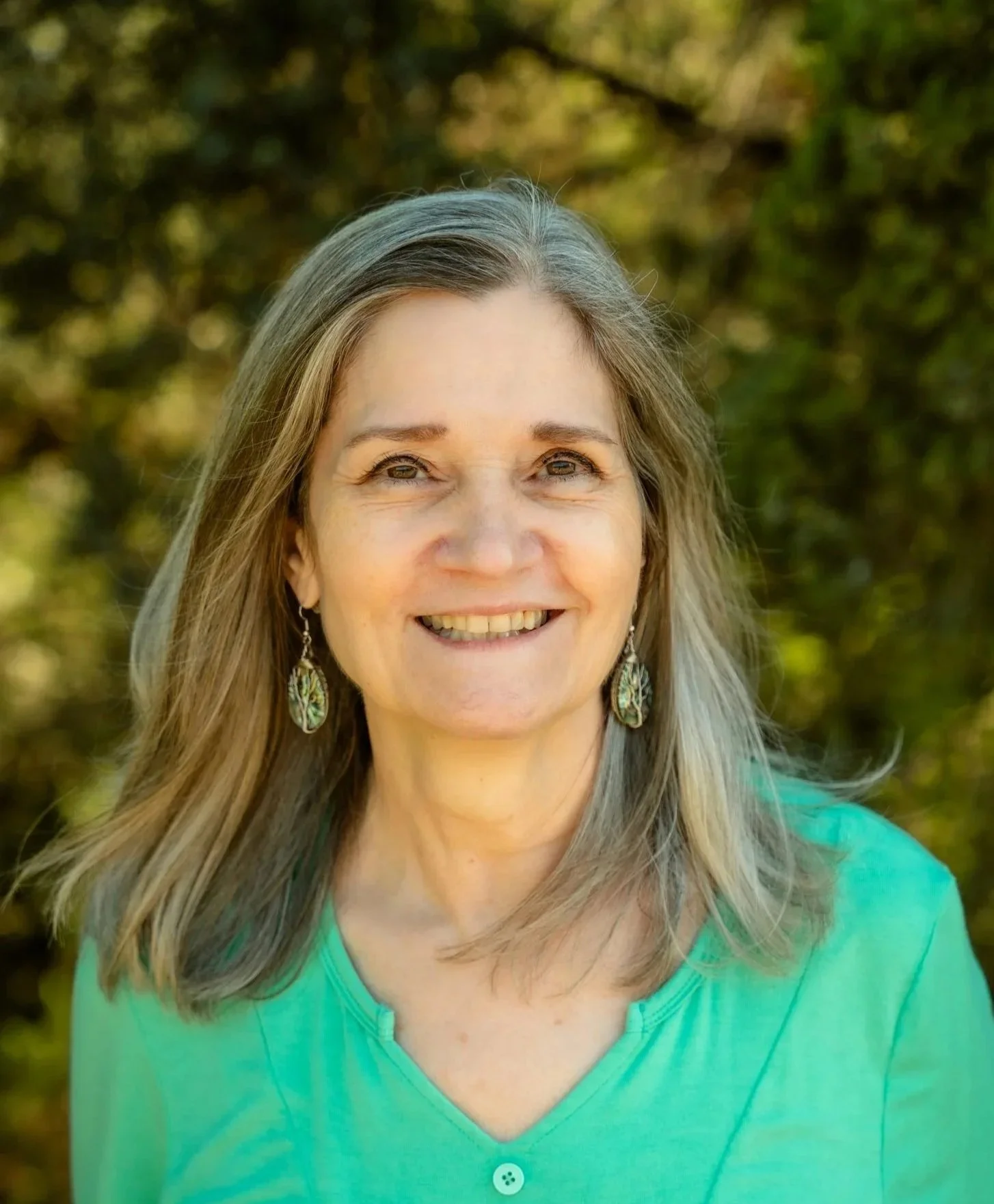 A smiling woman with long gray hair, wearing a green blouse and colorful earrings, standing outdoors with a blurred background of trees.