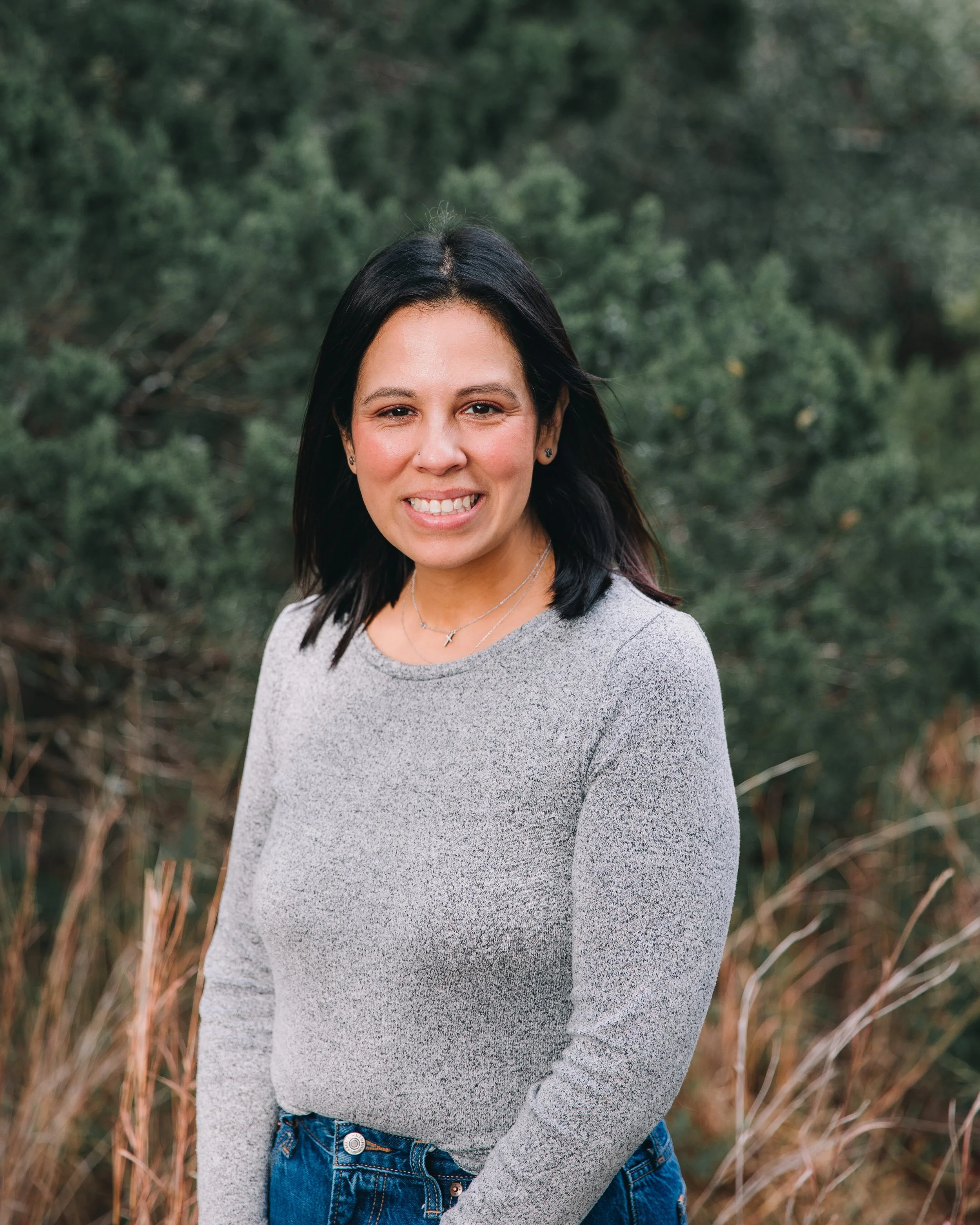 Portrait of a woman with shoulder-length dark hair and a grey top smiling outdoors with greenery in the background.