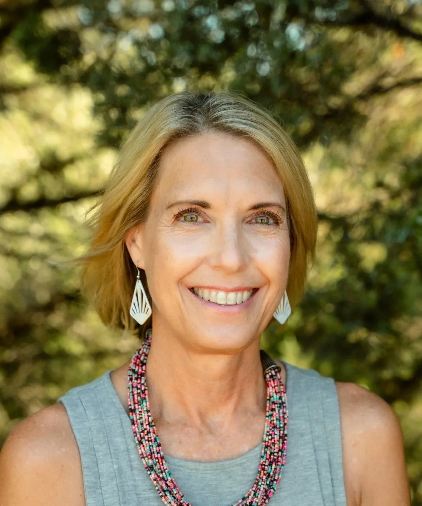 A smiling woman with short blonde hair, wearing a sleeveless gray top, colorful bead necklace, and white geometric earrings, standing outdoors with a background of greenery and trees.