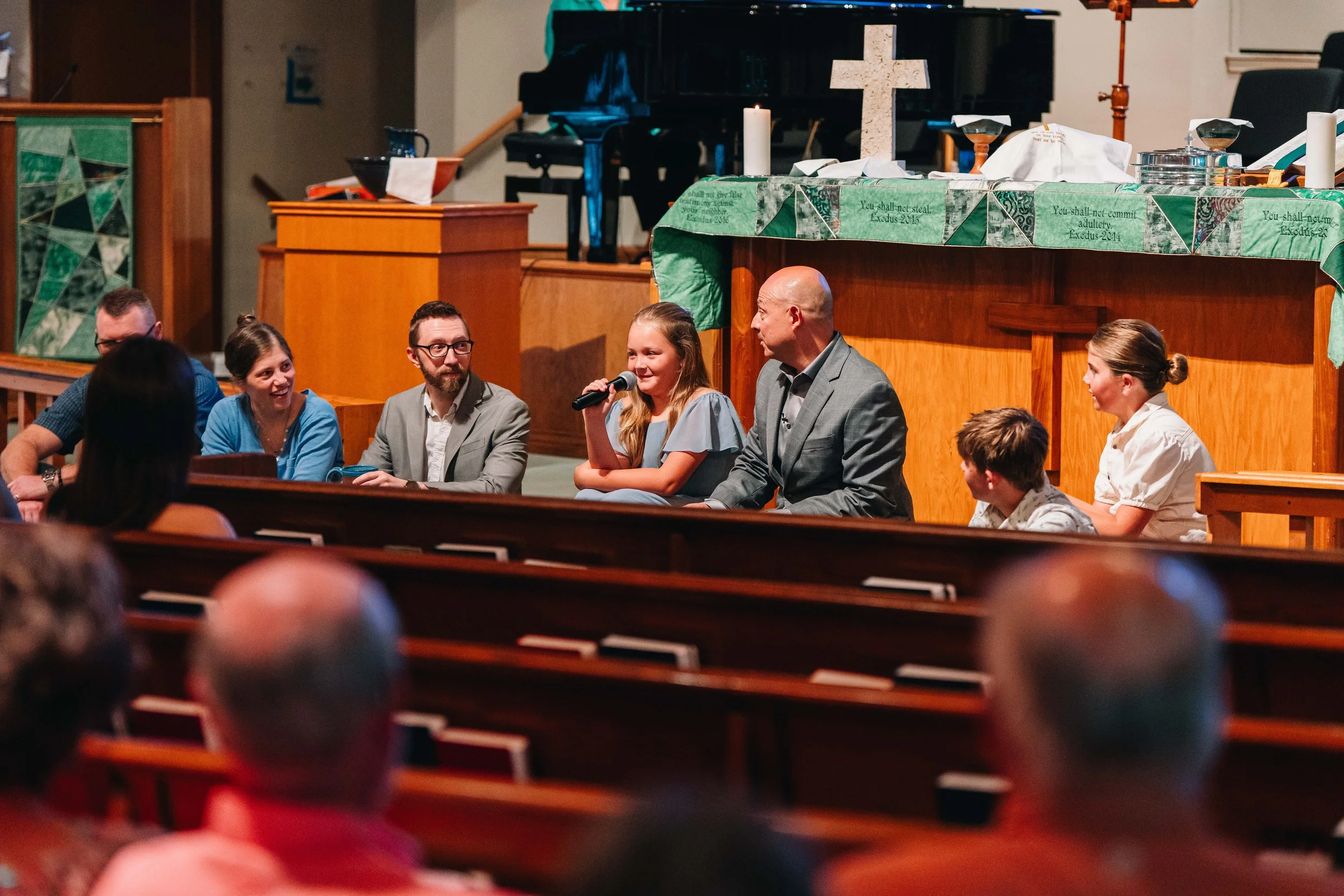 A group of people standing on the stage of a church, with a stained glass cross in the background, during a religious service or event, with congregation members seated in the pews facing the stage.