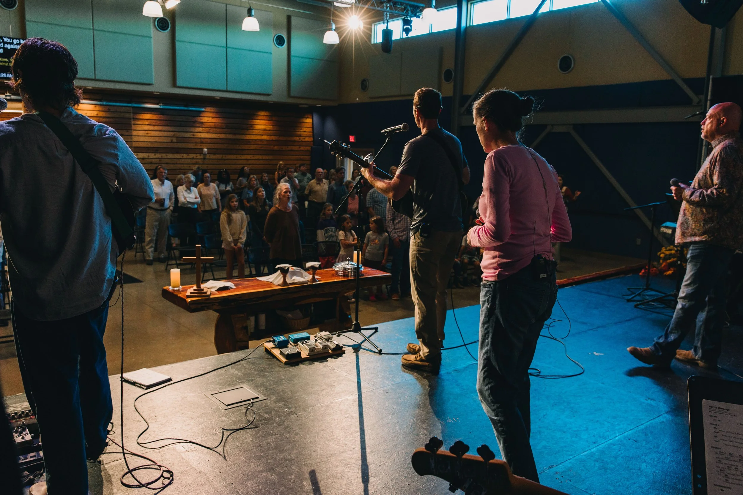 A church band performs on stage with a large cross on the left, a screen displaying lyric 'Great are you Lord' in the background, and an audience watching.