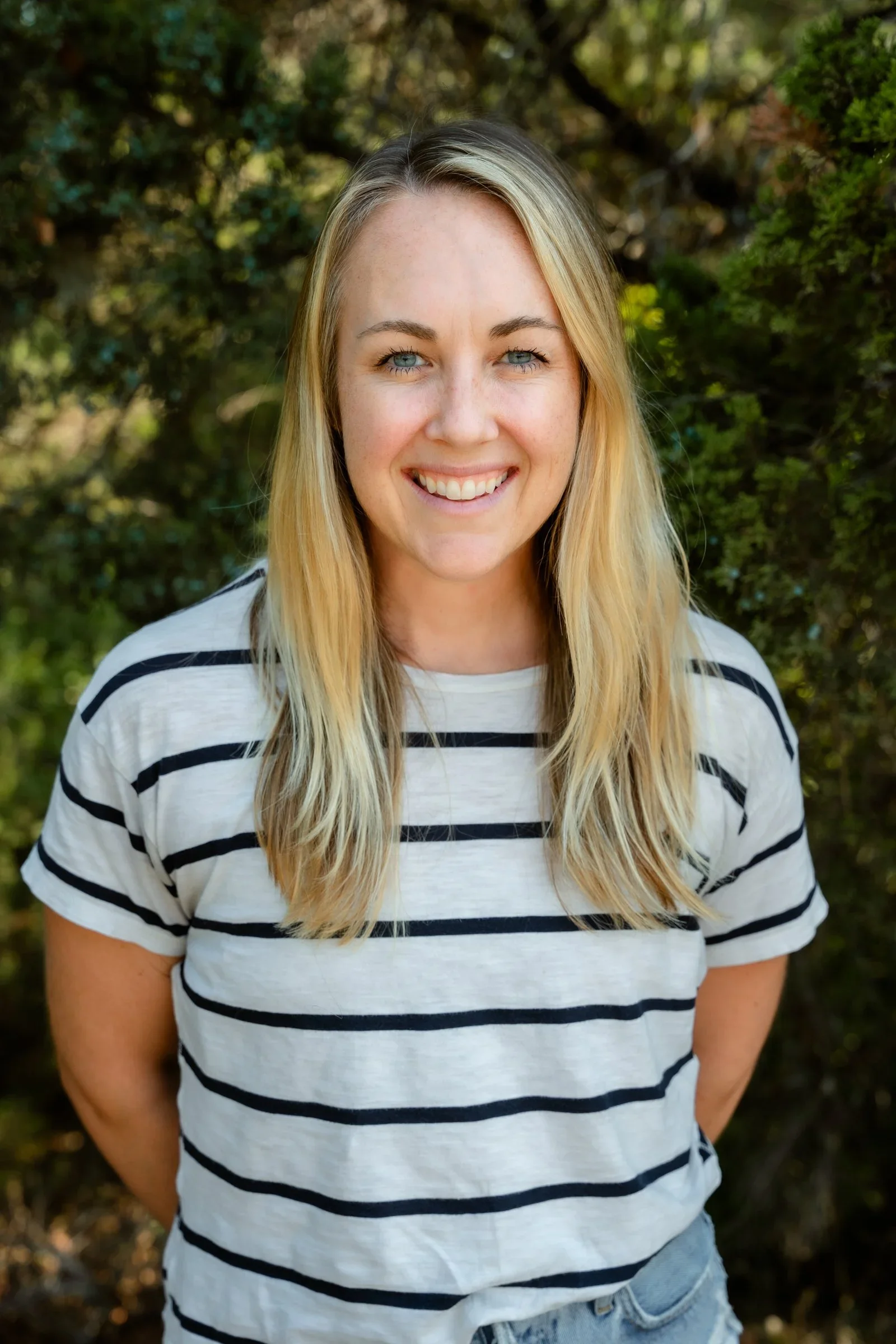 Young woman with blonde, shoulder-length hair styled in loose waves, smiling, wearing a beige t-shirt, gold earrings, and layered necklaces, standing outdoors with green foliage in the background.