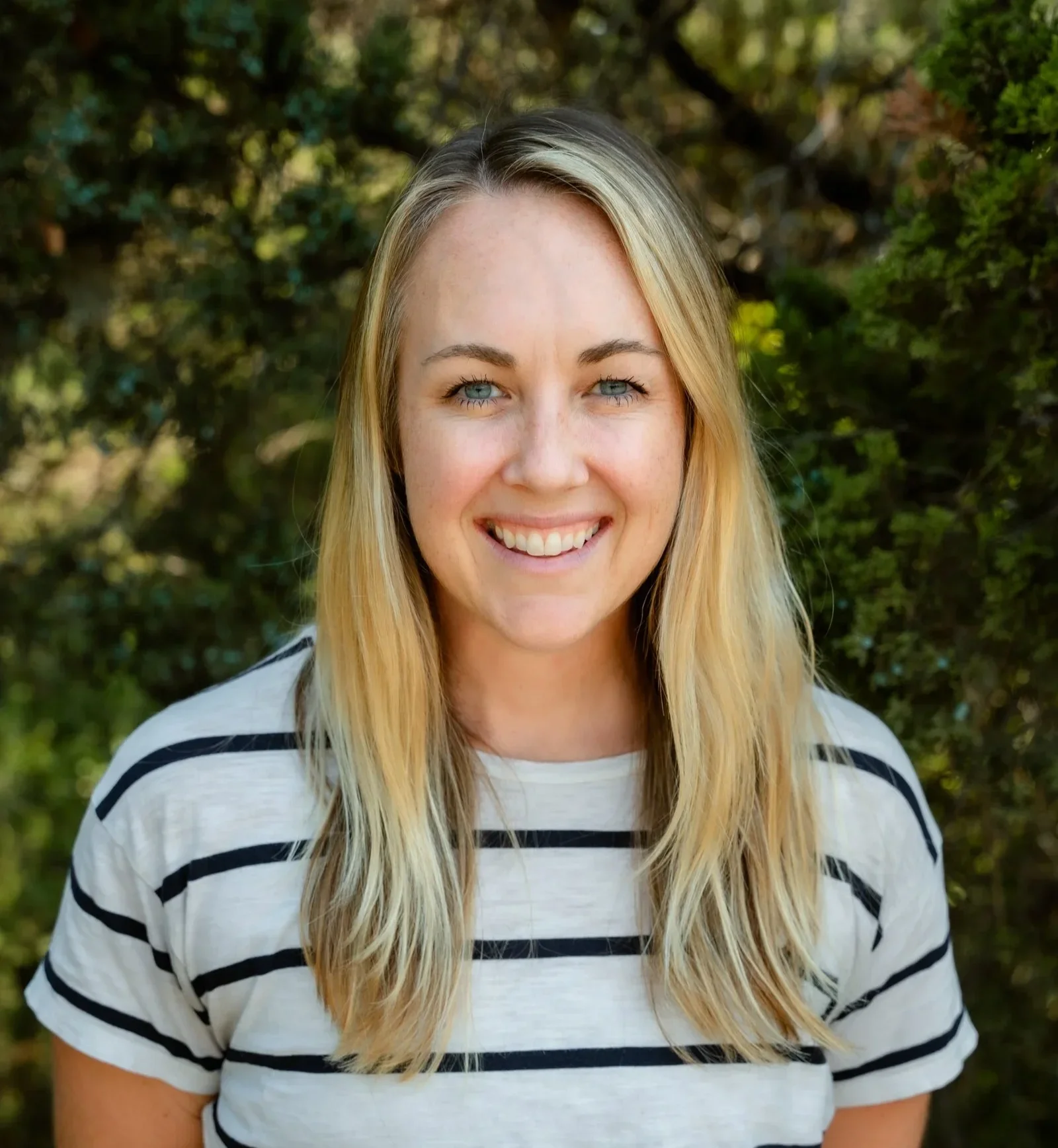 Young woman with blonde, shoulder-length hair styled in loose waves, smiling, wearing a beige t-shirt, gold earrings, and layered necklaces, standing outdoors with green foliage in the background.