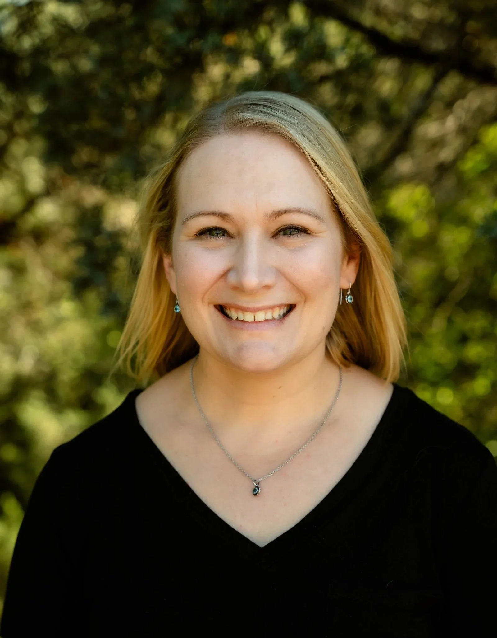 A young woman with blonde hair wearing a black top and jewelry, smiling outdoors with blurred green foliage background.