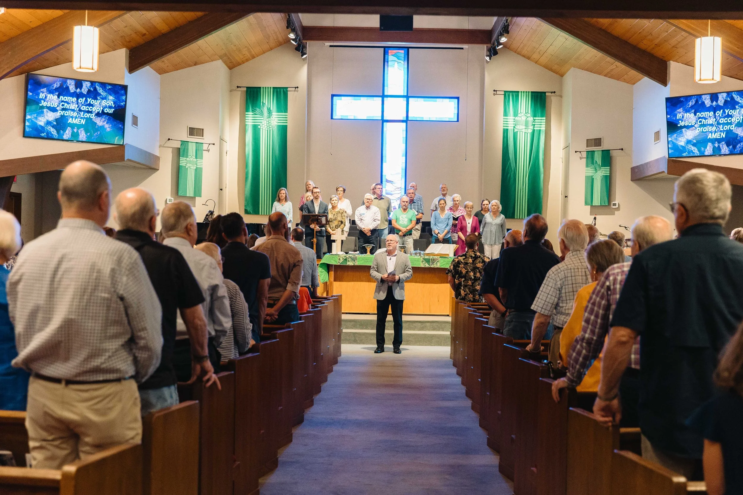 A group of people standing on the stage of a church, with a stained glass cross in the background, during a religious service or event, with congregation members seated in the pews facing the stage.