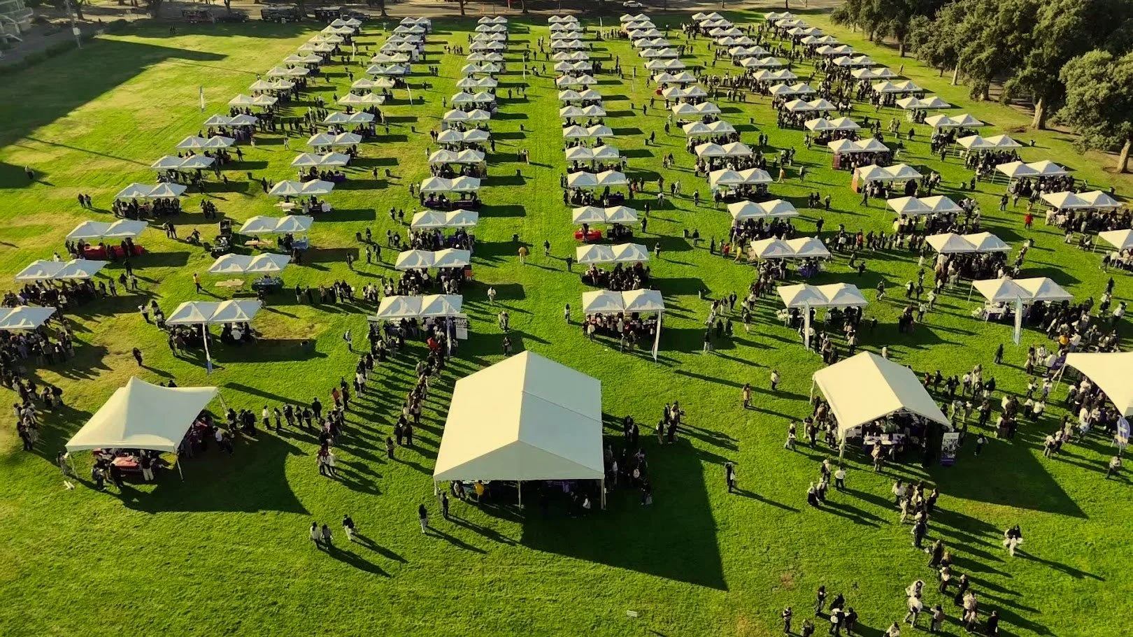 Event with multiple white canopy tents arranged across a large outdoor field with guests attending a festival or corporate gathering.