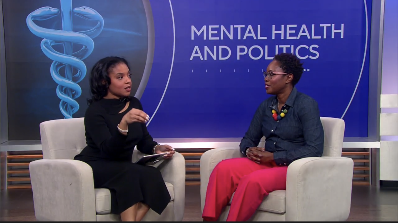 A woman in professional attire standing in a television news studio with a large screen displaying the logo 'LNC Truth' and multiple monitors in the background.