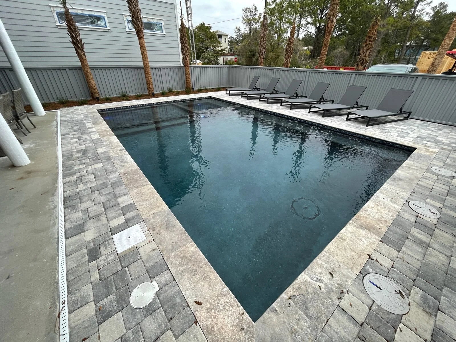 A backyard with a rectangular swimming pool surrounded by gray paving stones. Lounge chairs are lined up along the back fence, and tall palm trees are visible behind the fence. A gray house with three windows is in the background.