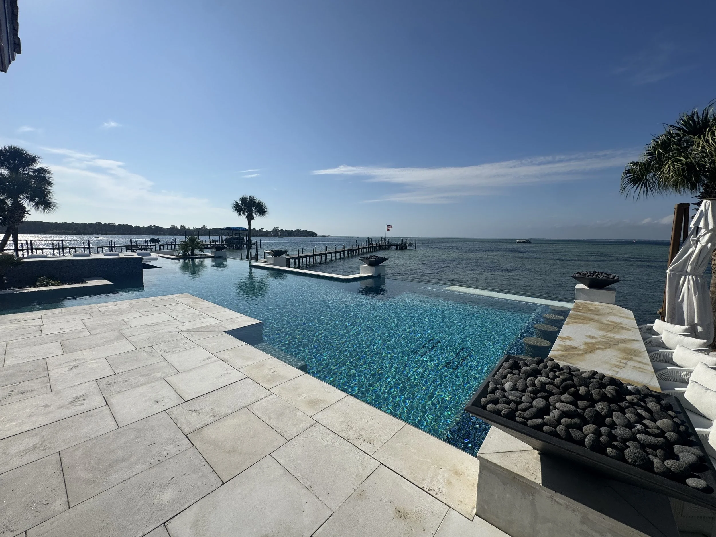 Pool with ocean view, palm trees, and a wooden pier extending into the water on a clear, sunny day.