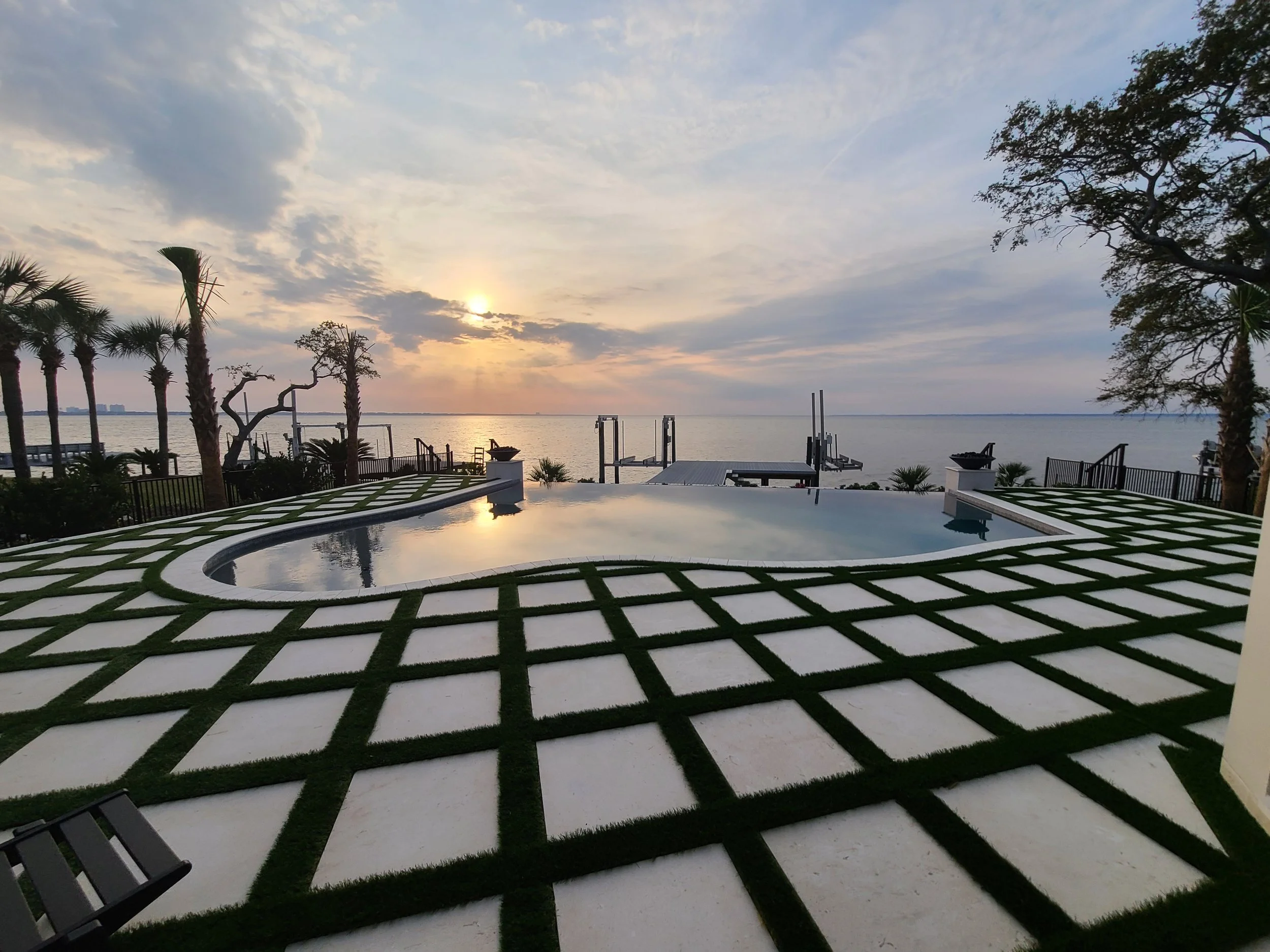 A view of a waterfront at sunset with palm trees, a swimming pool, and outdoor seating, overlooking a body of water with a cloudy sky.