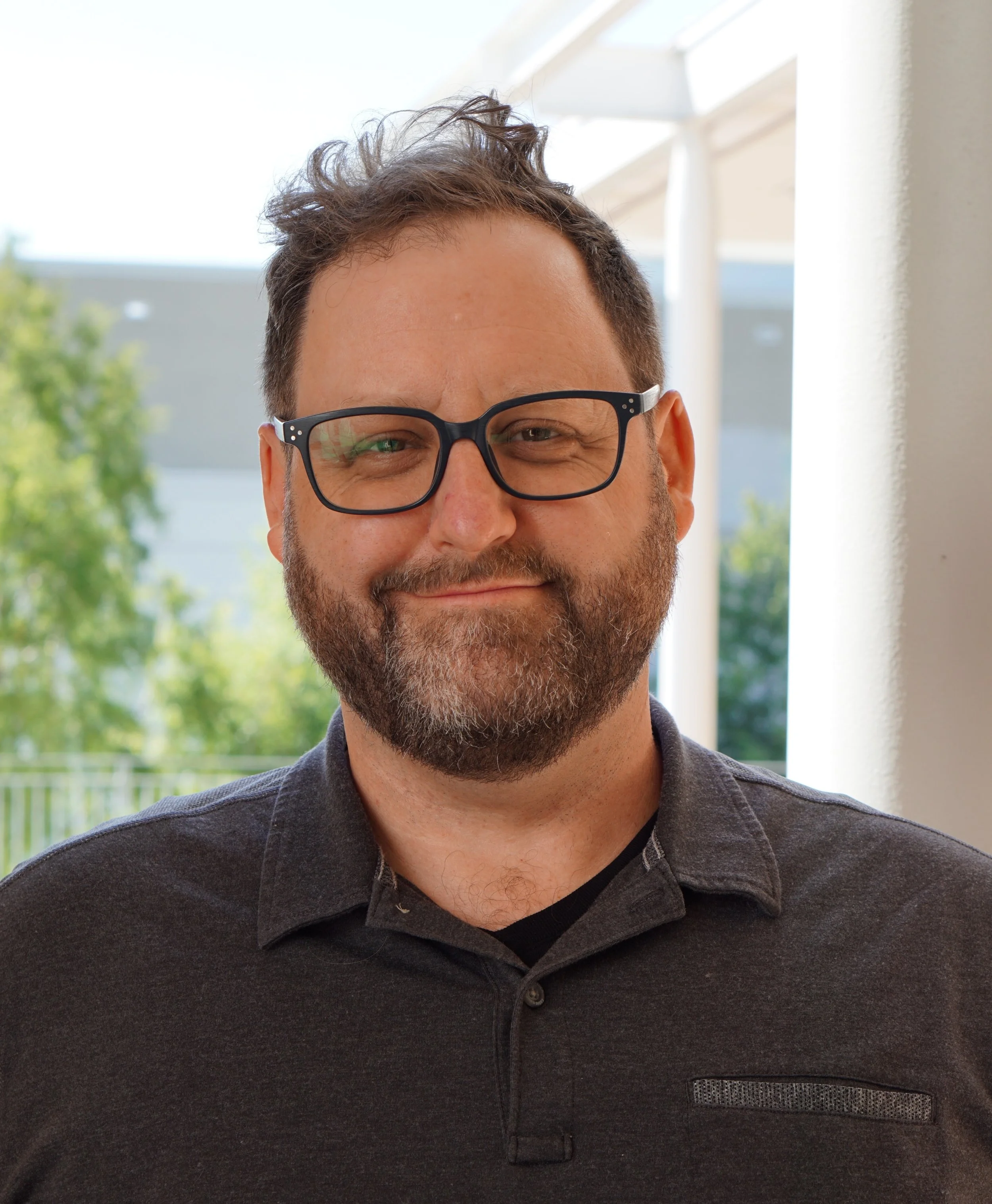 Portrait of a man with glasses, beard, and mustache, wearing a navy blue vest over a plaid shirt, smiling indoors.