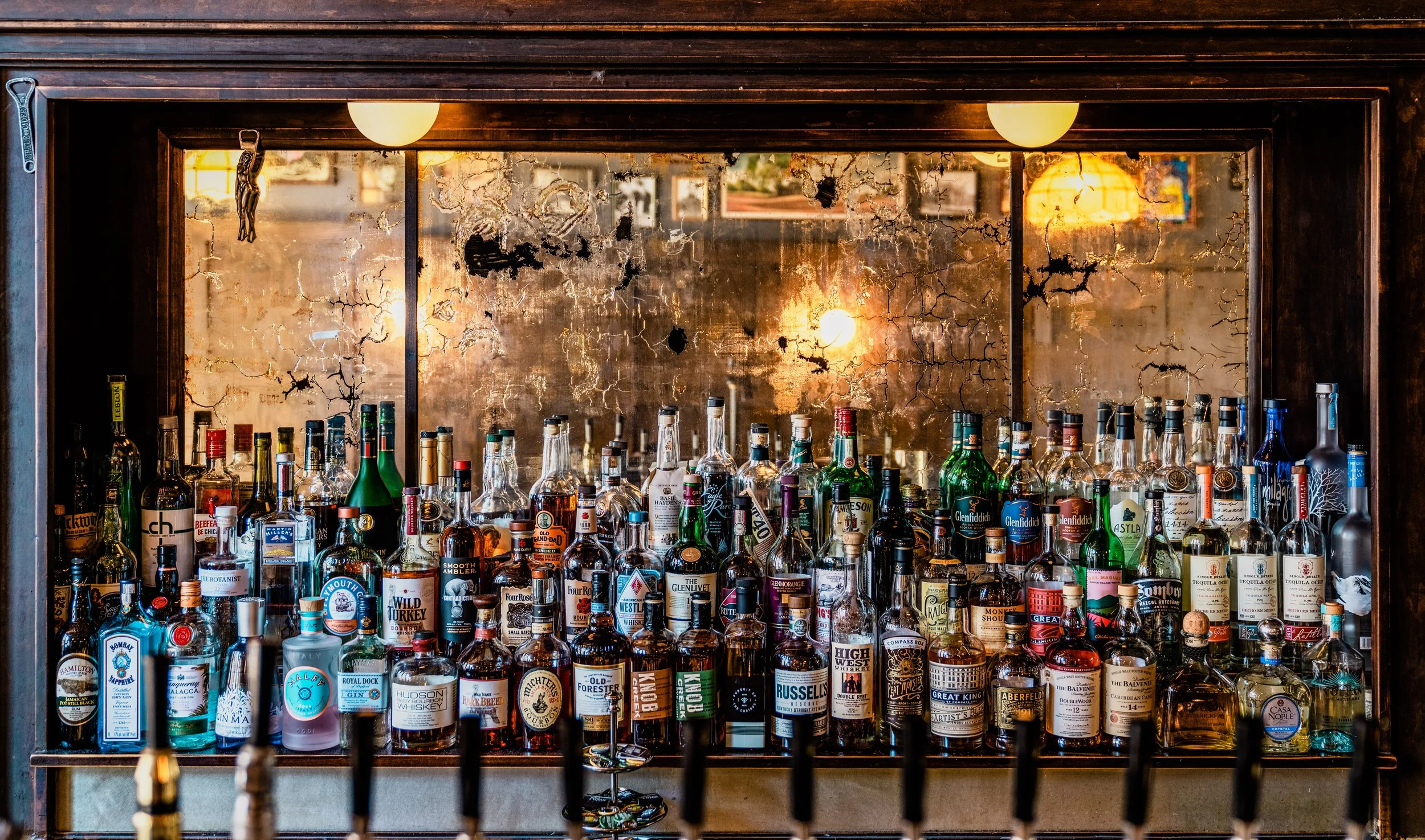 The Swill Inn by Siren Betty Design, mirrored back bar with antique patina and rows of colorful liquor bottles.
